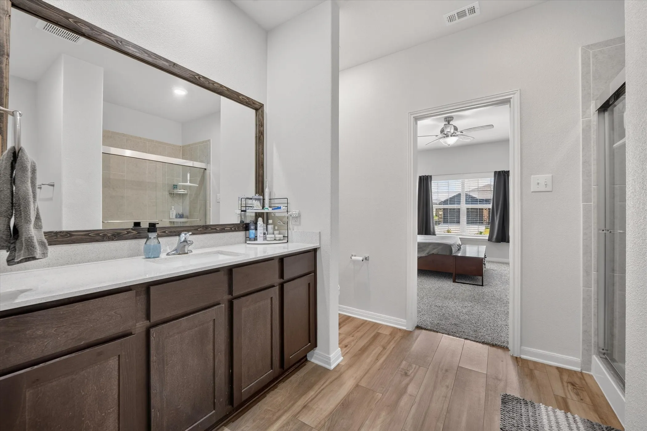 Double vanity with white countertop, wood-finish flooring, framed mirror, and glass-enclosed shower