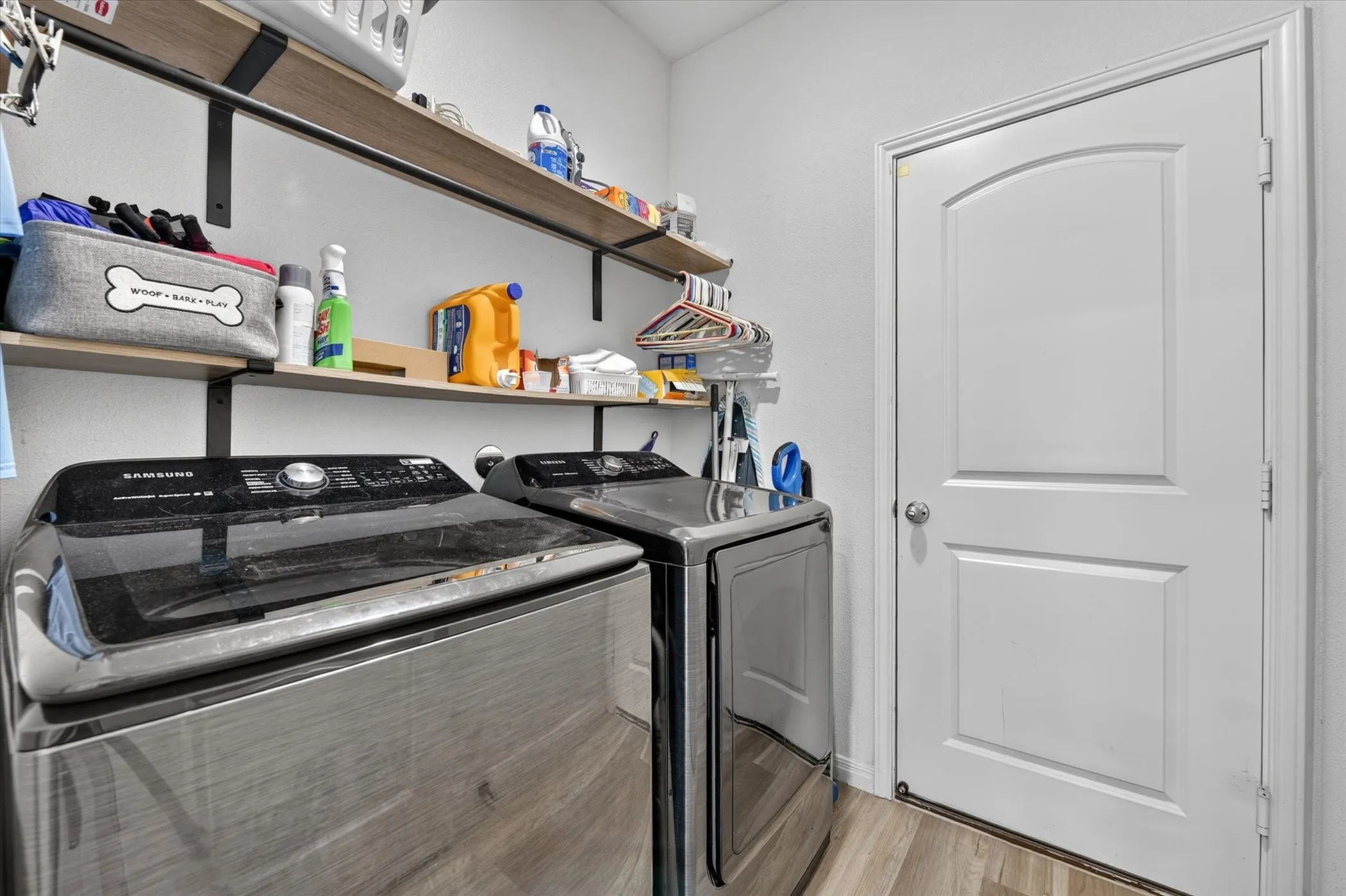 Laundry area featuring wood-finish flooring, two large appliances, two wall-mounted shelves, and a paneled door