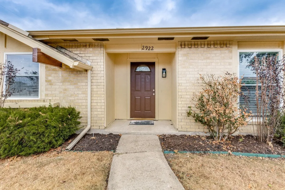 Brick exterior with a covered entry, dark wood-finish front door, exterior lighting, and mature landscaping