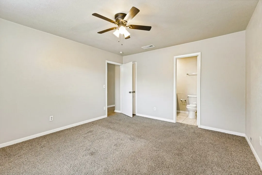 Carpeted room with light-toned walls, ceiling fan, and adjacent bathroom featuring a toilet and tile flooring