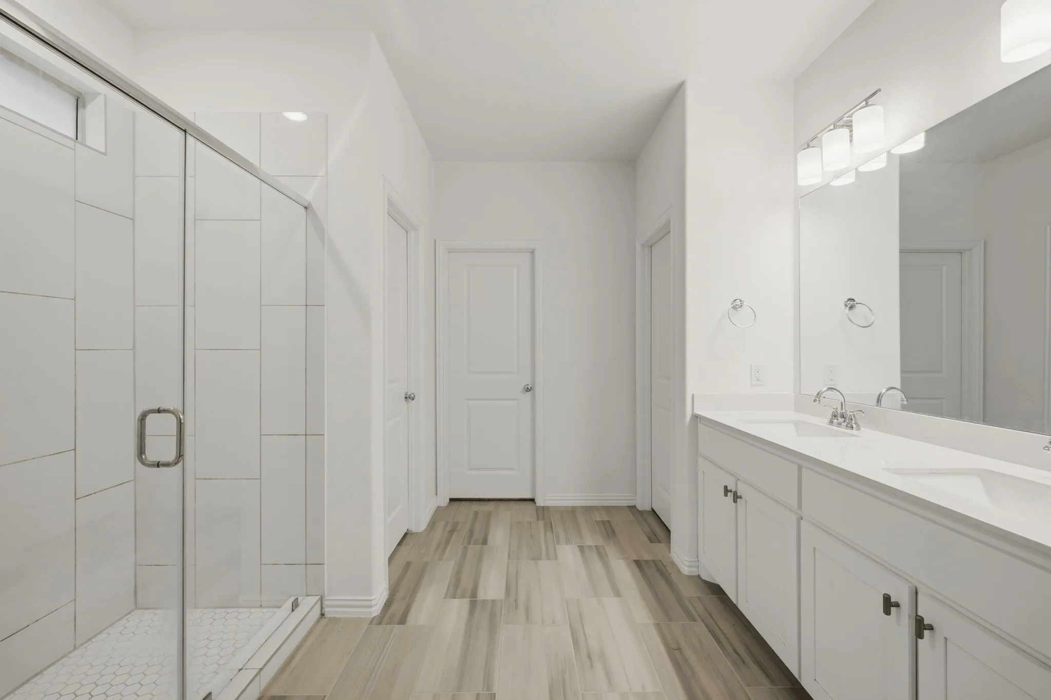 Spacious bathroom featuring a glass-enclosed shower with subway tile, dual vanity with undermount sinks, wood-finish flooring, and white cabinetry