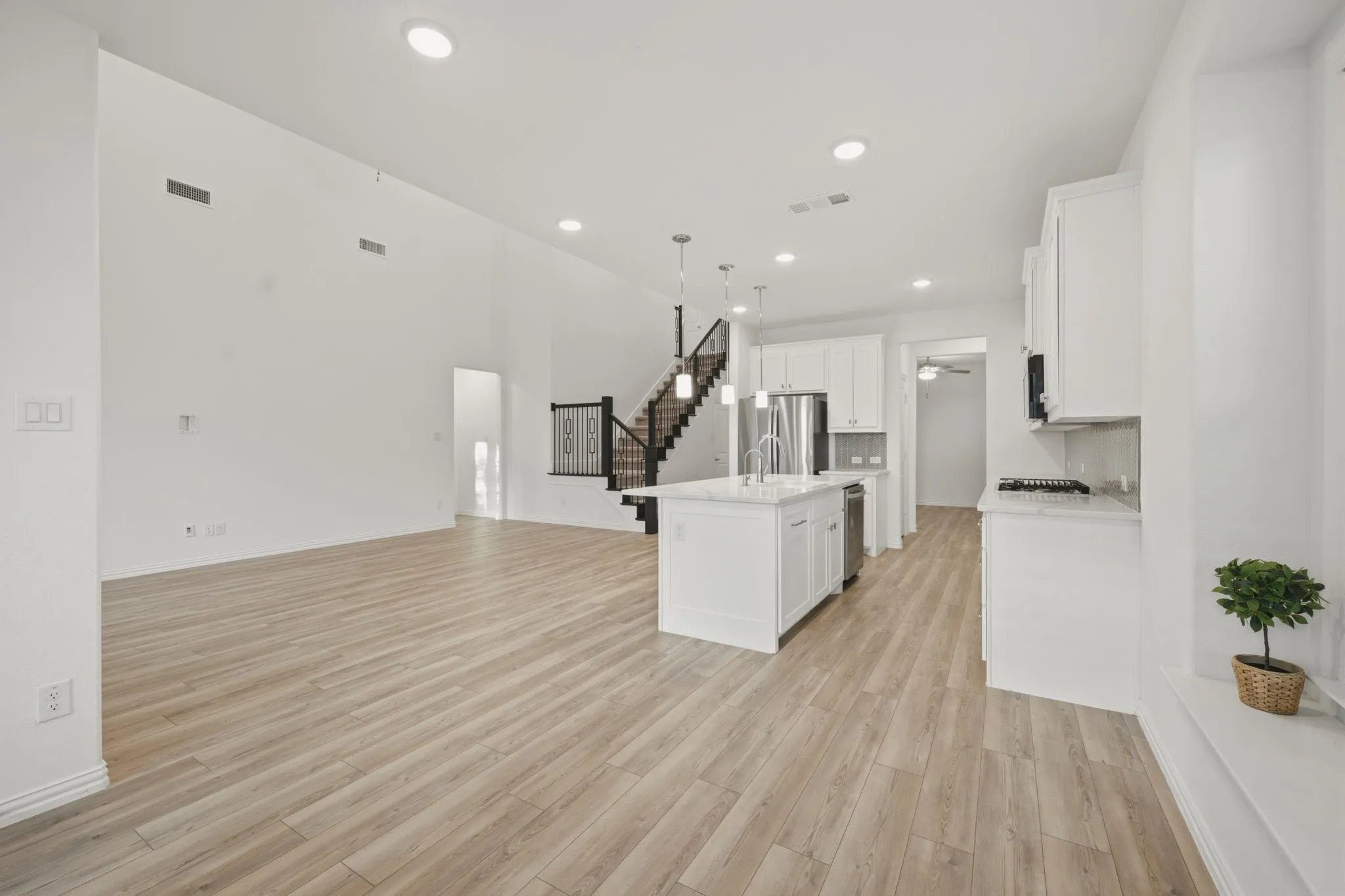 Open-concept living area featuring wood-finish flooring, recessed lighting, a modern kitchen island with pendant lighting, stainless steel appliances, and a staircase with wrought iron balusters