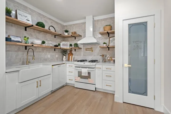 Kitchen featuring light wood-finish flooring, white cabinetry with gold-tone hardware, a white farmhouse sink with a gold-tone faucet, a gas range with a matching range hood, and open wood shelving