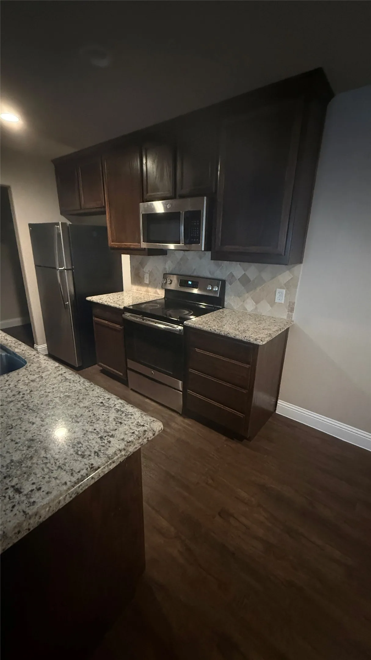 Kitchen featuring dark wood cabinetry, stainless steel appliances, granite countertops, a tiled backsplash, and wood-finish flooring