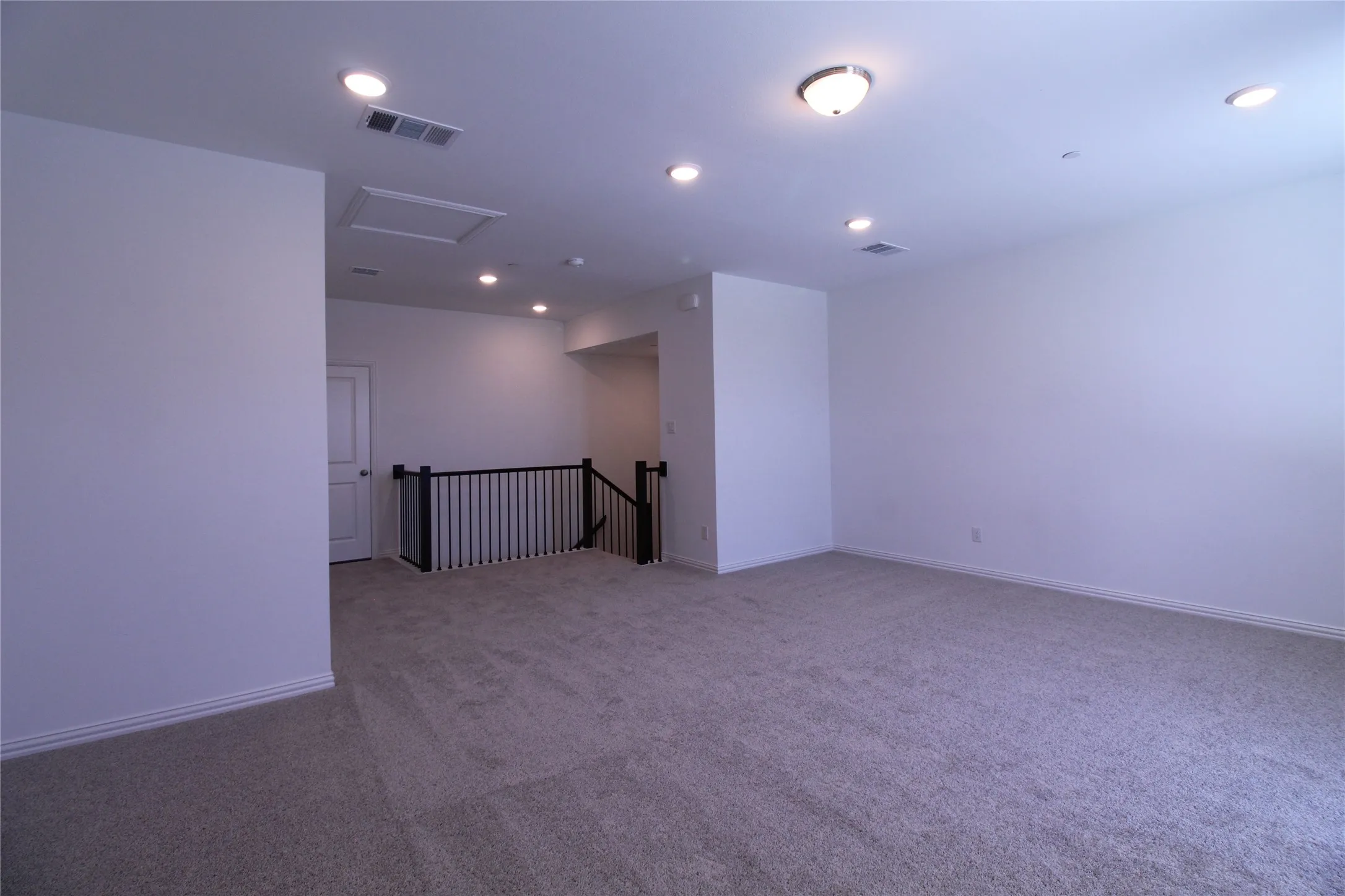Spacious carpeted area featuring a prominent black spindle railing, white baseboards, recessed ceiling lighting, and a white interior door