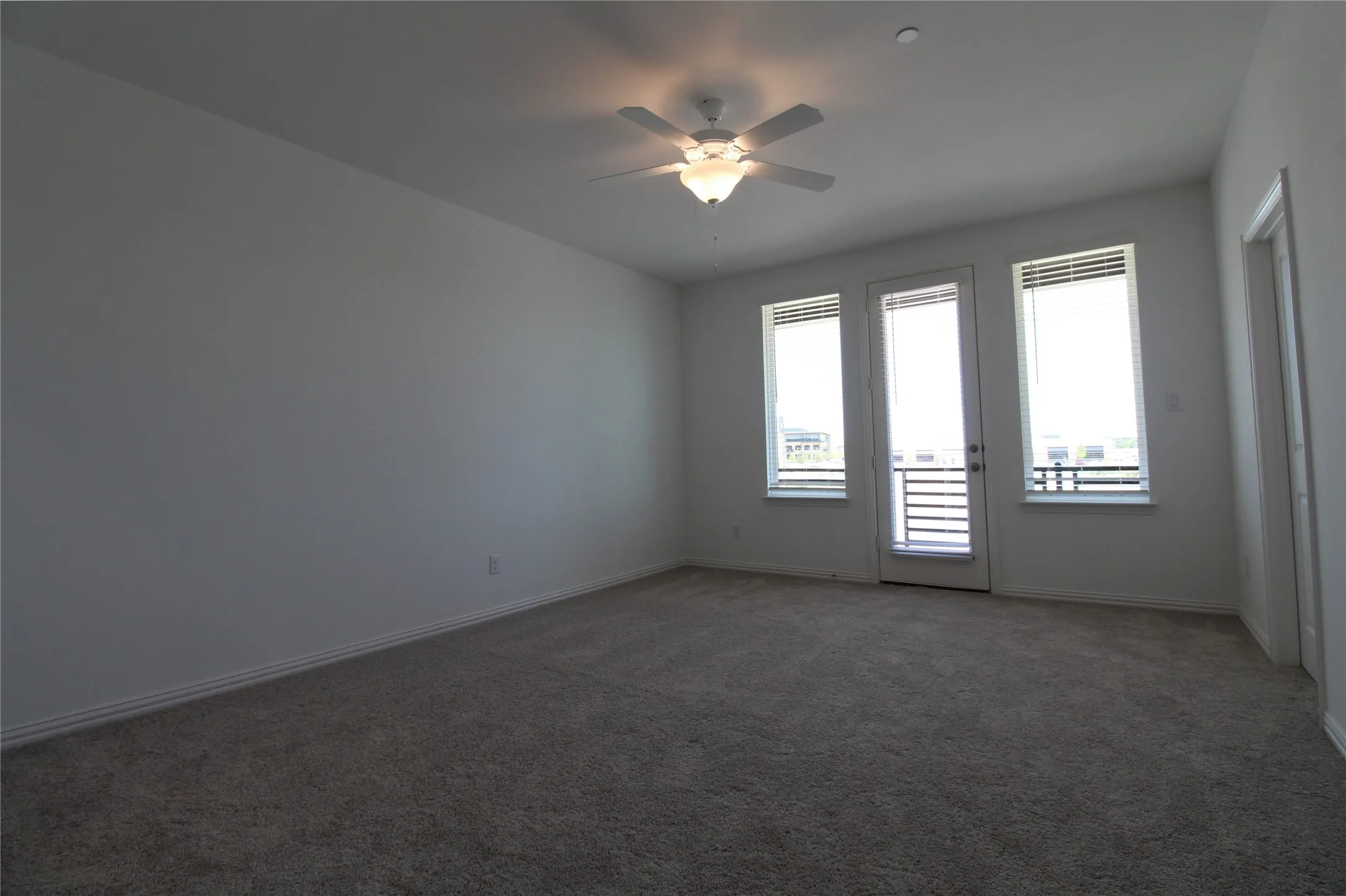 Spacious room featuring carpeting, white walls, a ceiling fan with integrated lighting, and a door leading to a balcony flanked by two windows with horizontal blinds
