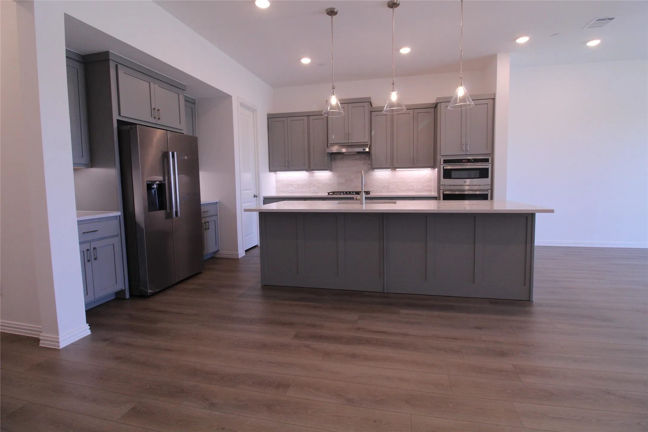 Open-concept kitchen featuring an oversized island with pendant lighting, shaker-style cabinetry, stainless steel appliances, and wood-finish flooring