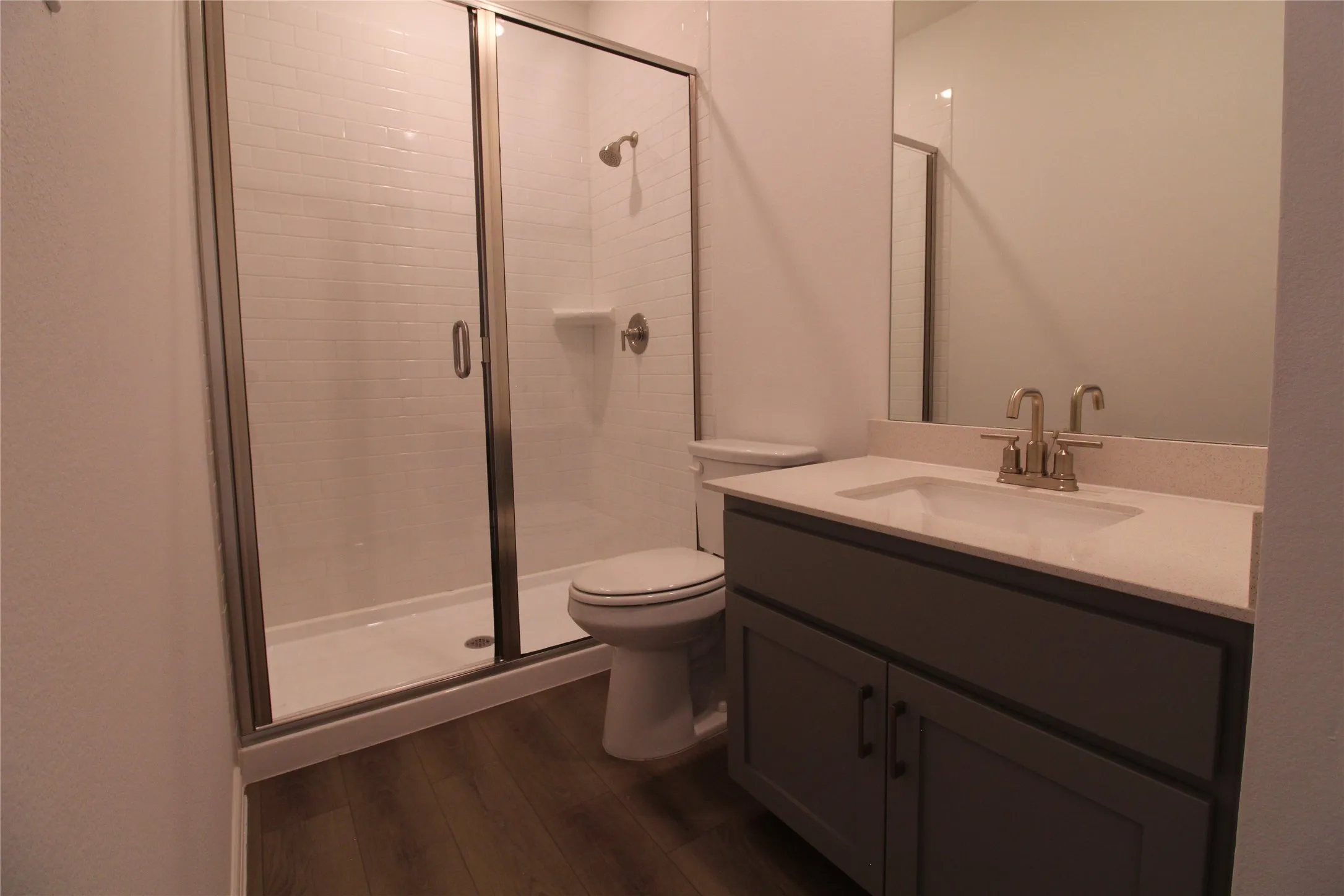 Bathroom featuring a walk-in shower with subway tile surround and glass enclosure, a single vanity with a light-colored countertop and undermount sink, brushed nickel fixtures, and wood-finish flooring