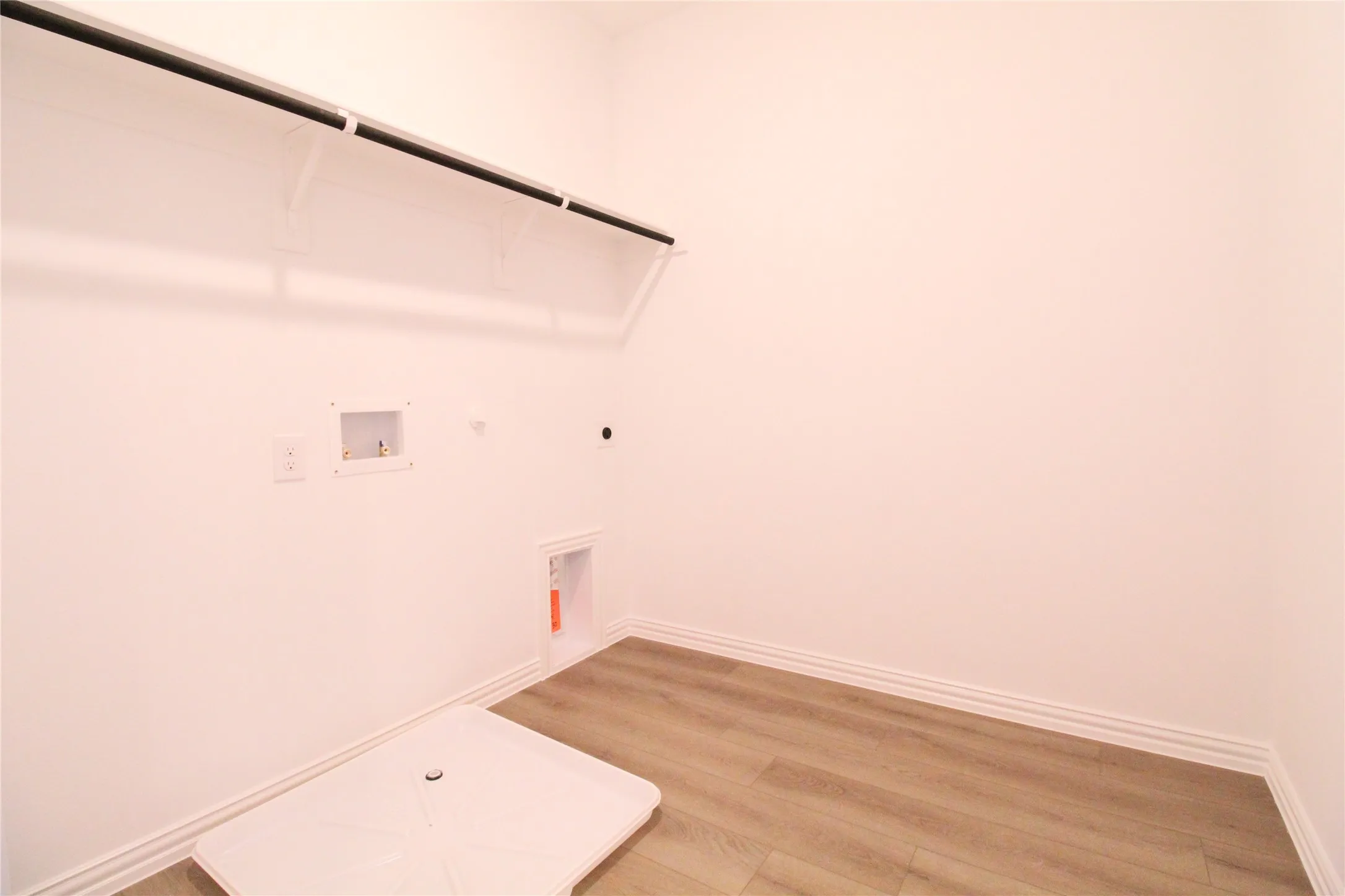 Utility space featuring wood-finish flooring, white baseboards, a white storage shelf with a black rod, and appliance hookups