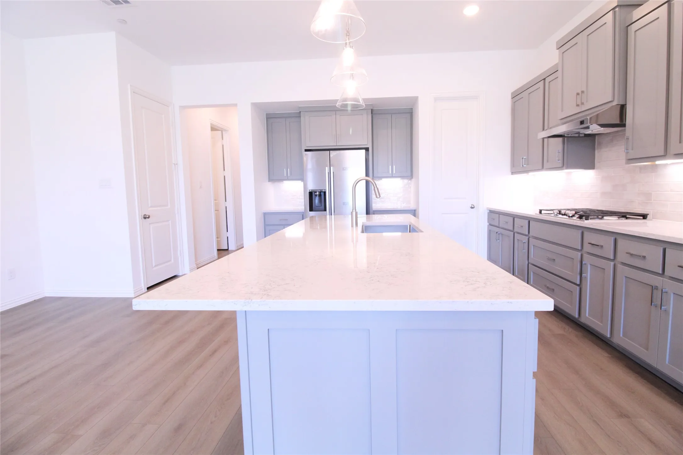 Kitchen featuring a large central island with a built-in sink and stainless steel faucet, light gray cabinetry, white subway tile backsplash, stainless steel appliances, and wood-finish flooring
