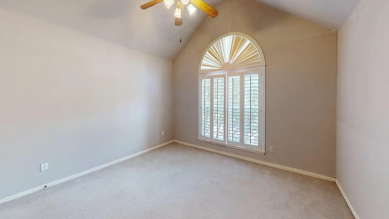Vaulted ceiling room featuring light-colored carpeting, a large arched window with plantation shutters, and a ceiling fan with integrated lighting