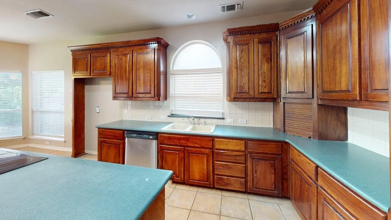 Spacious kitchen featuring extensive wood cabinetry with decorative crown molding, turquoise quartz countertops, tile flooring, and abundant natural light from an arched window