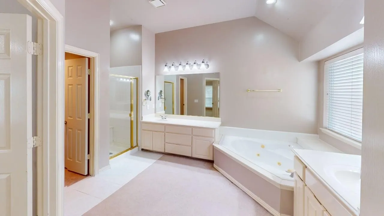 Vaulted ceiling, two vanities with integrated sinks, corner jetted tub, glass-enclosed shower, and window with blinds