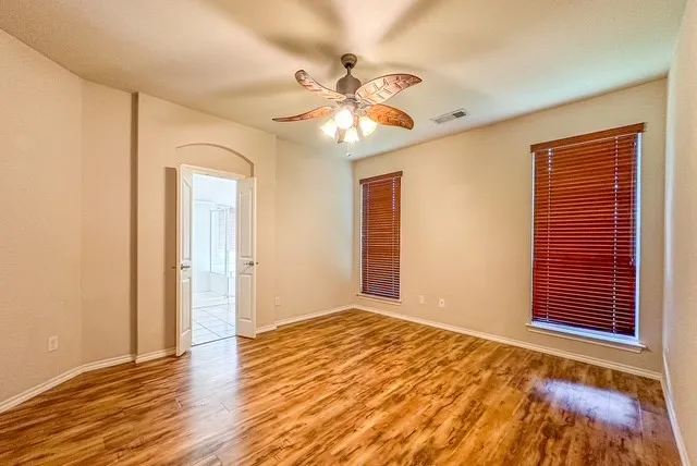 Wood-finish flooring, ceiling fan with integrated lighting, two windows with wood blinds, neutral wall tones, and an arched doorway