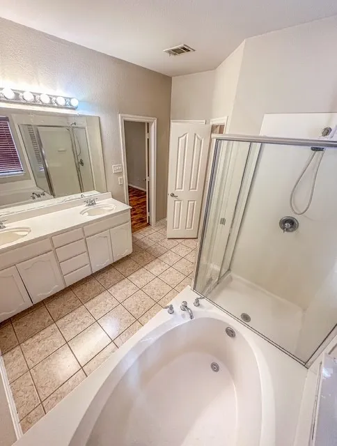 Dual vanity with integrated sinks, full-width mirror, tile flooring, built-in soaking tub, and enclosed shower