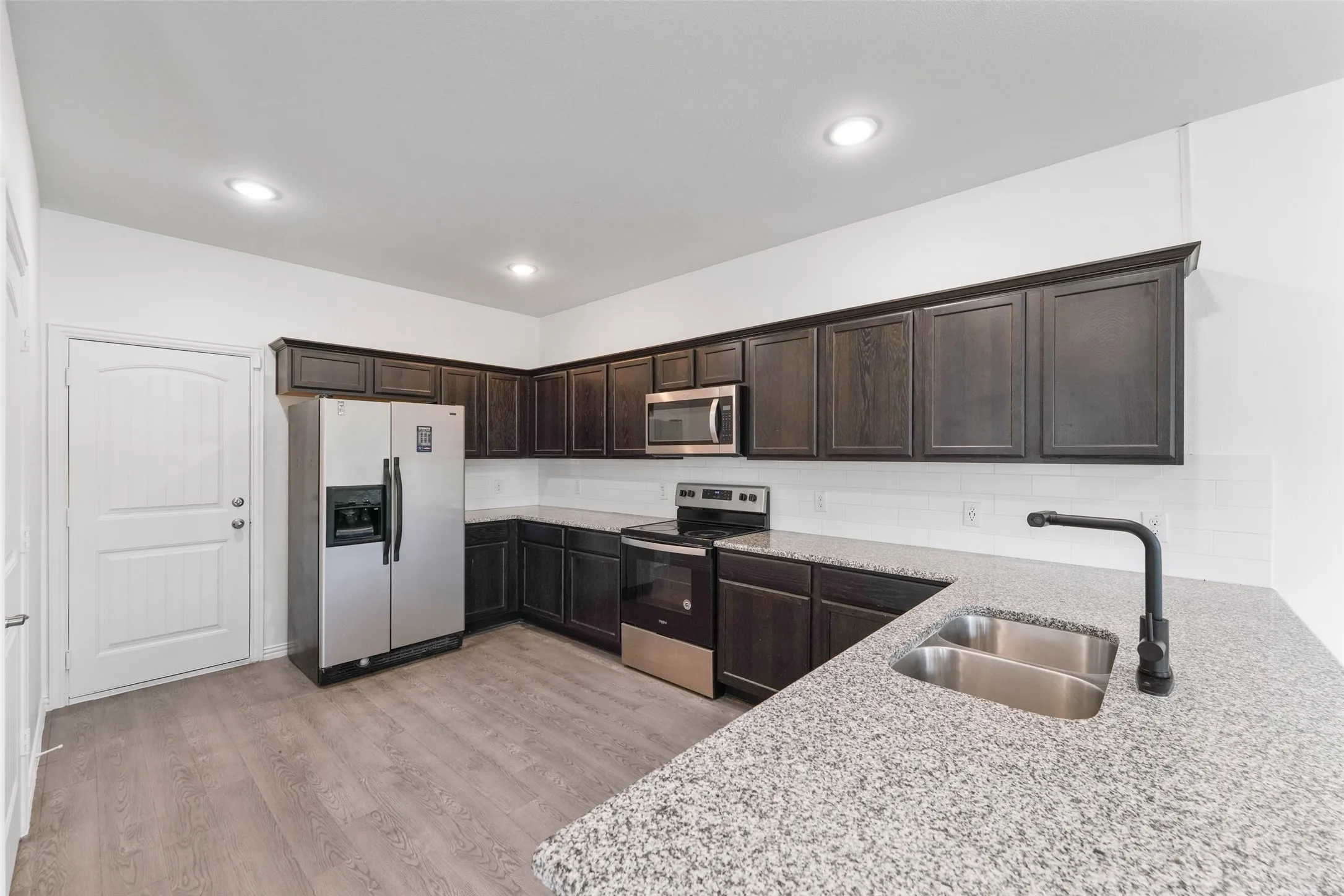 Modern kitchen featuring dark wood cabinetry with glass-front upper doors, stainless steel appliances including a refrigerator and range, quartz countertops with waterfall edge, a deep stainless steel sink with matte black pull-down faucet, recessed ceiling lighting, and wood-finish flooring throughout the open layout
