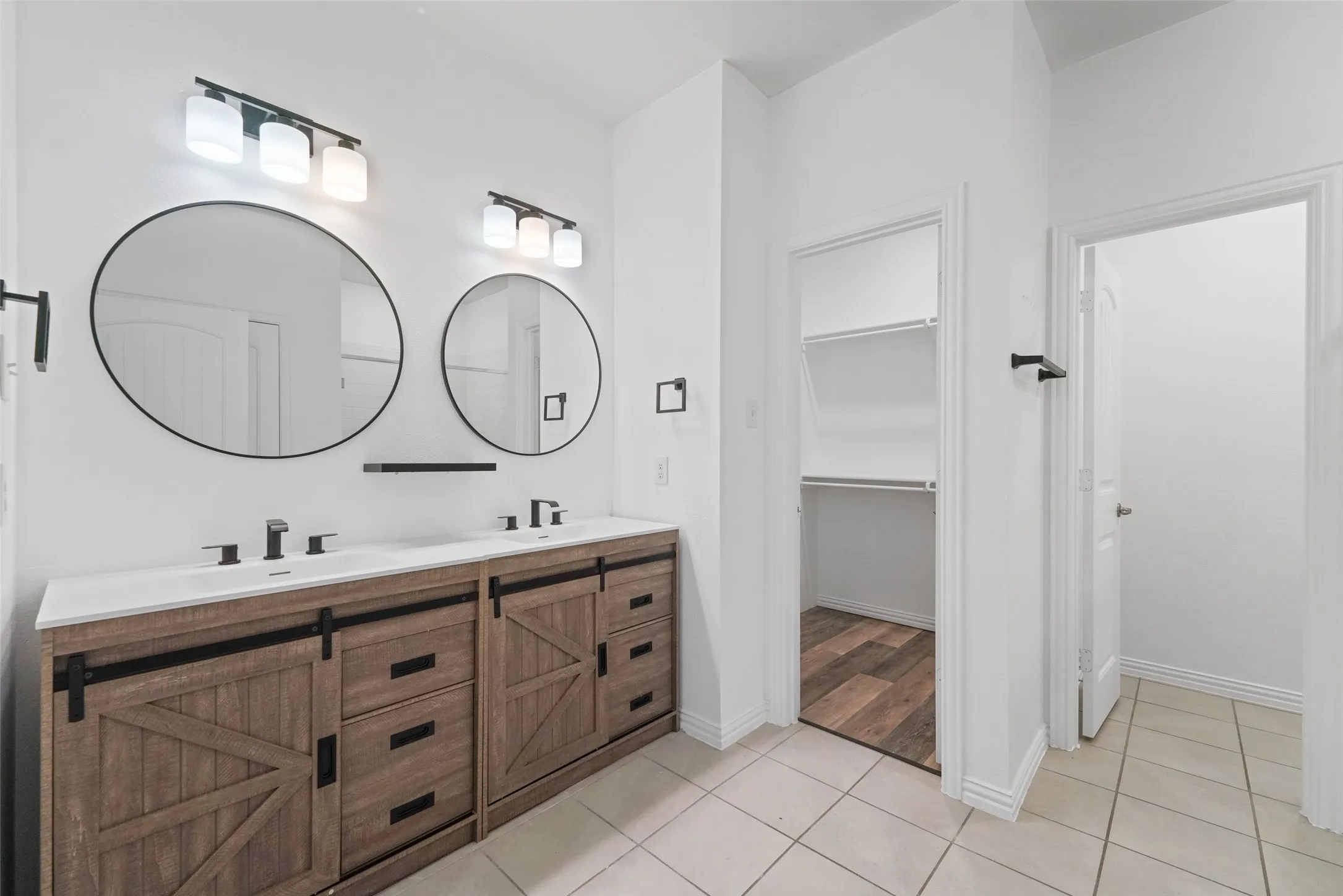 Spa-inspired primary bathroom showcasing a custom wood-finish dual vanity with black hardware and metal accents, paired round mirrors with integrated lighting, tile flooring, and a walk-in shower with frameless glass enclosure
