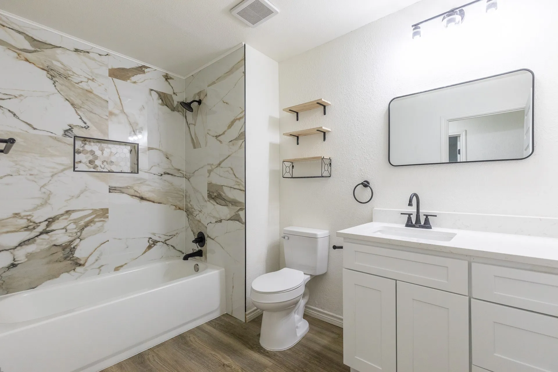 Bathroom featuring a white vanity with an integrated sink, black-framed mirror, wood-finish flooring, and a bathtub with a tiled surround