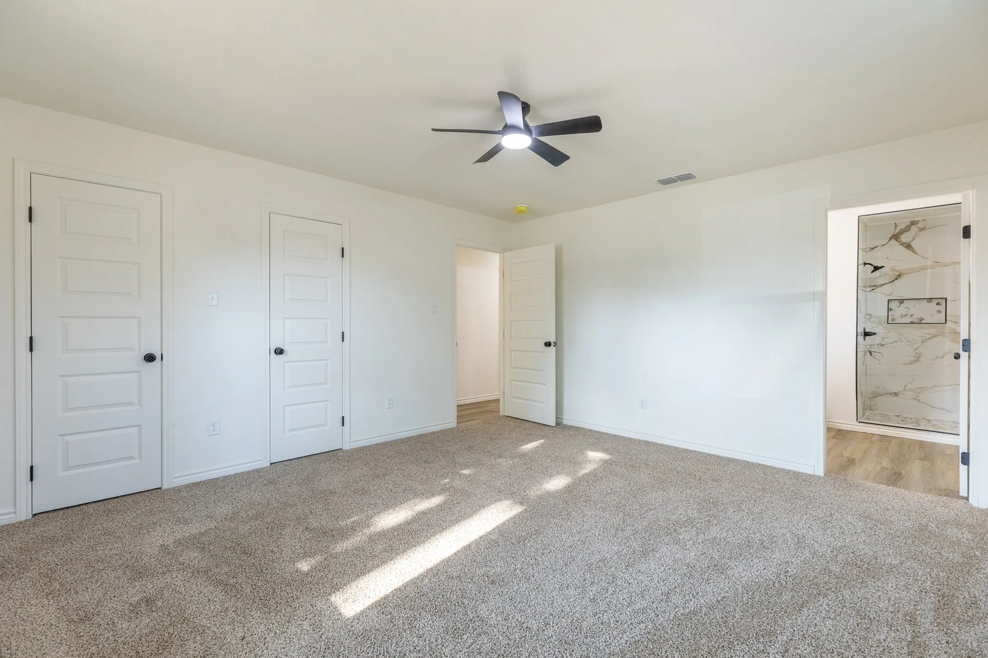 Spacious room with plush carpeting, a ceiling fan, two paneled doors, and access to a bathroom featuring a walk-in shower with tiled walls