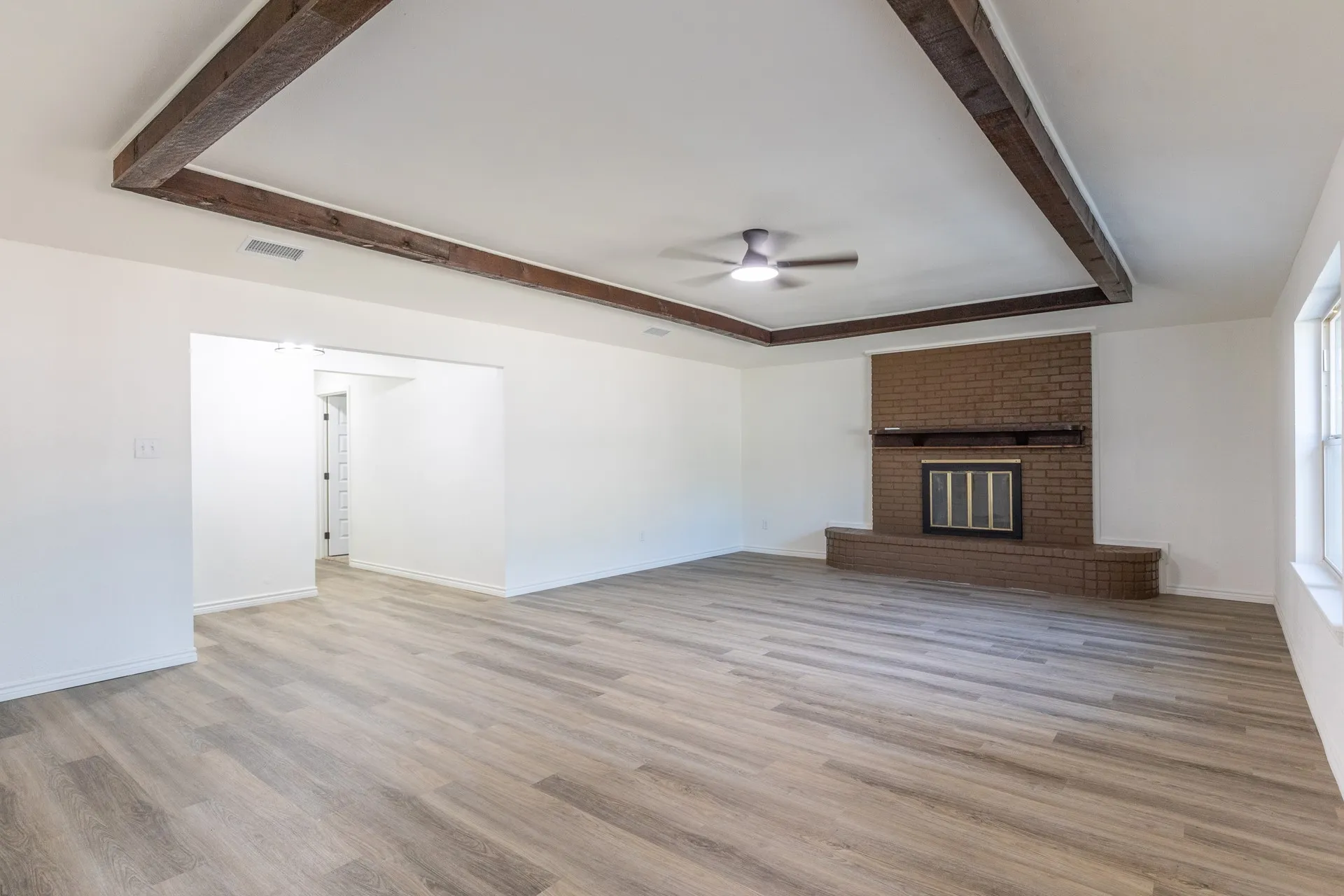Spacious room featuring wood-finish flooring, a tray ceiling with exposed wood beams, a ceiling fan, and a brick fireplace