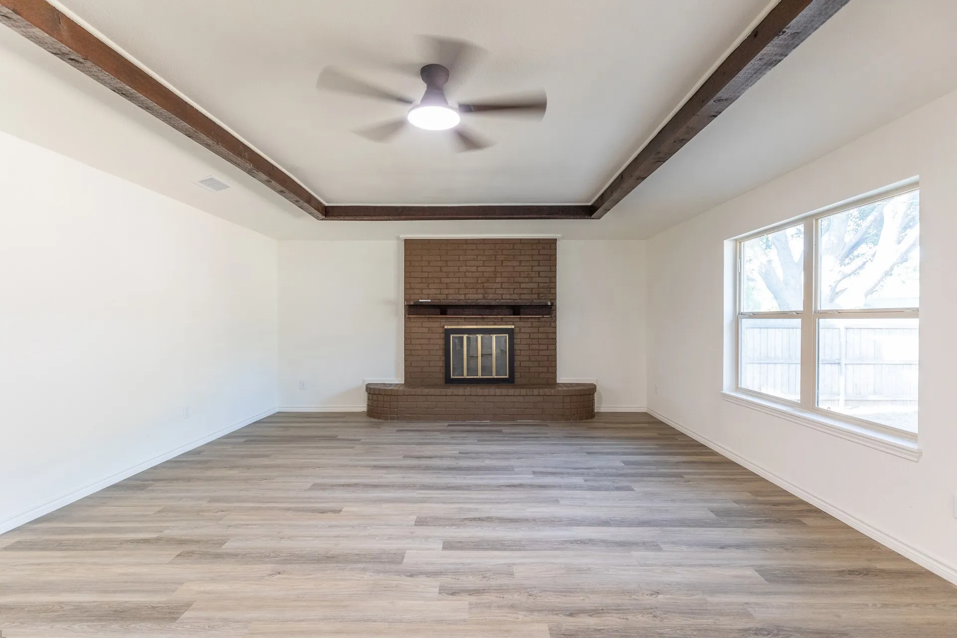Tray ceiling with wood-finish beams, wood-finish flooring, brick-clad fireplace with hearth, and a multi-pane window