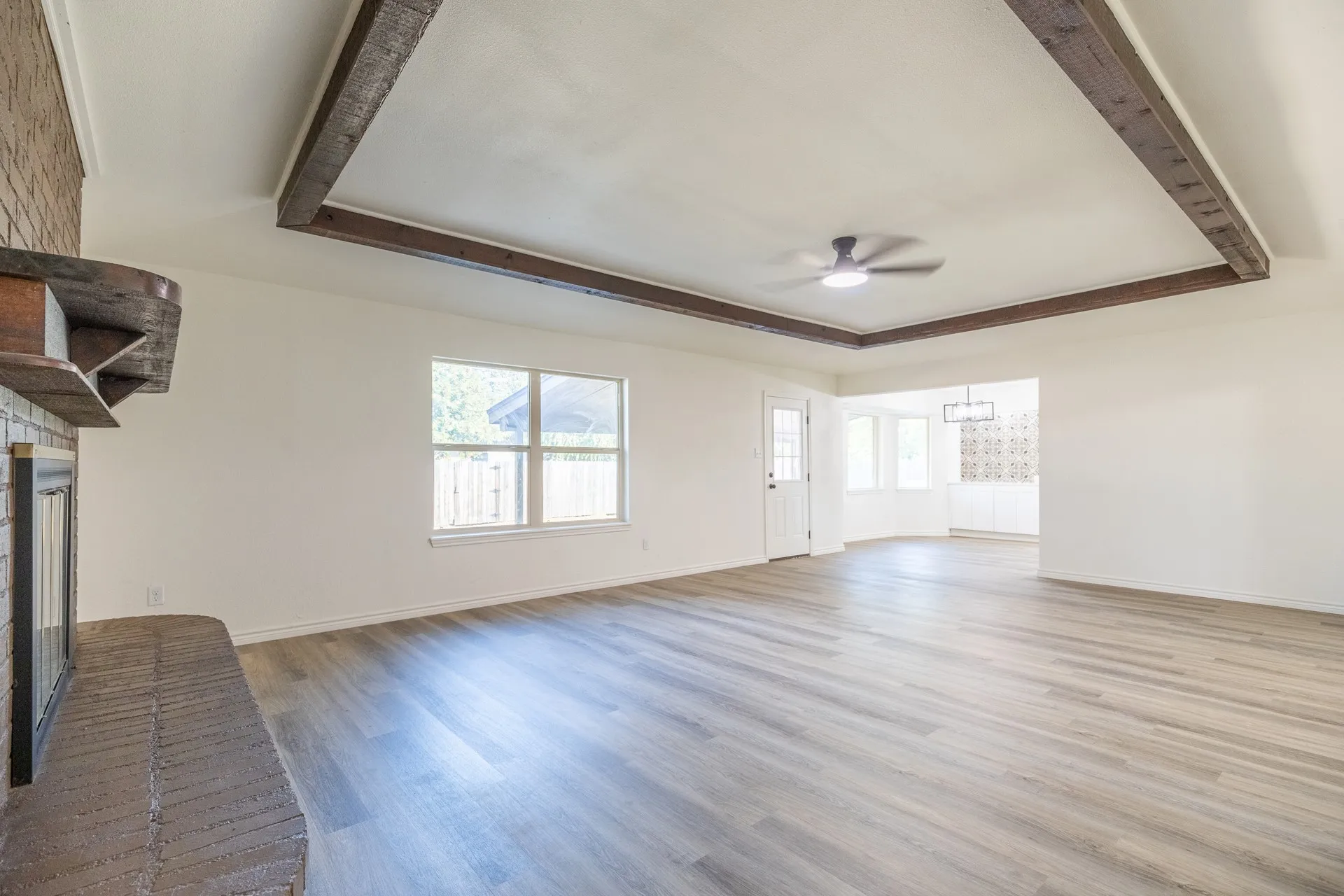 Tray ceiling with exposed wood beams, wood-finish flooring, brick fireplace with wood mantel, ceiling fan, and multiple windows