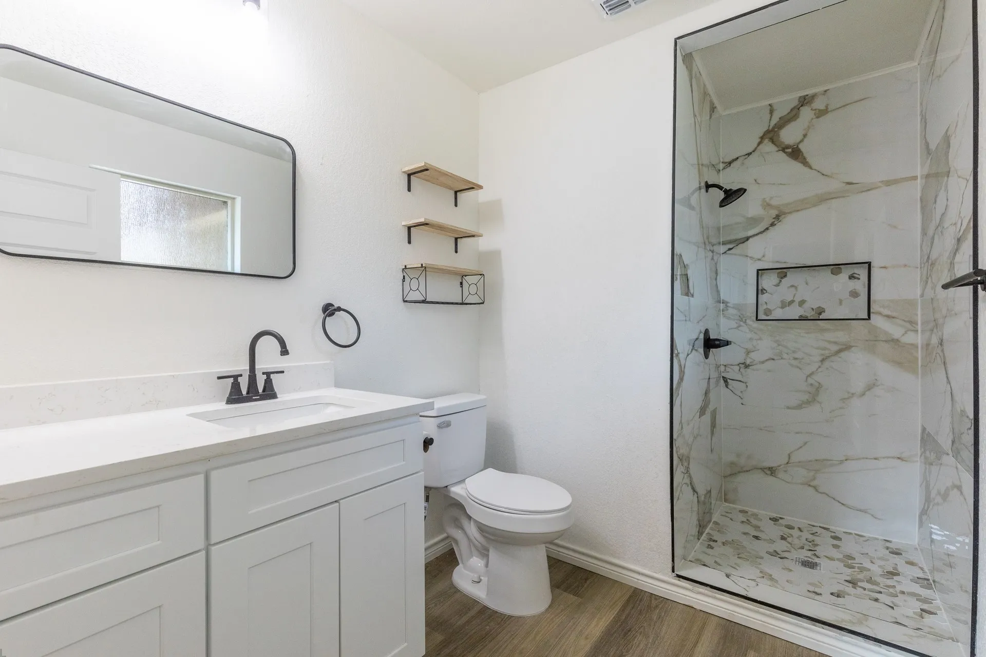 Bathroom featuring a white vanity with a white countertop, a black-framed mirror, a walk-in shower with stone-look tile, a shower niche, and wood-finish flooring