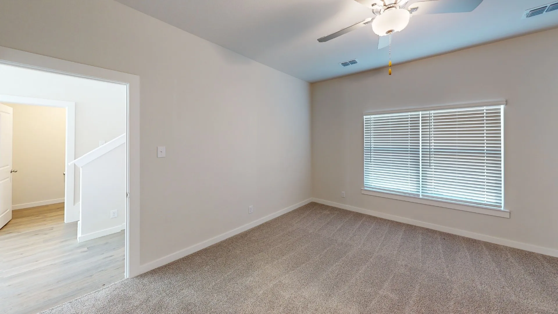 Neutral tone wall paint, carpeted flooring, window with horizontal blinds, overhead ceiling fan with light fixture, and wood-finish flooring visible through doorway