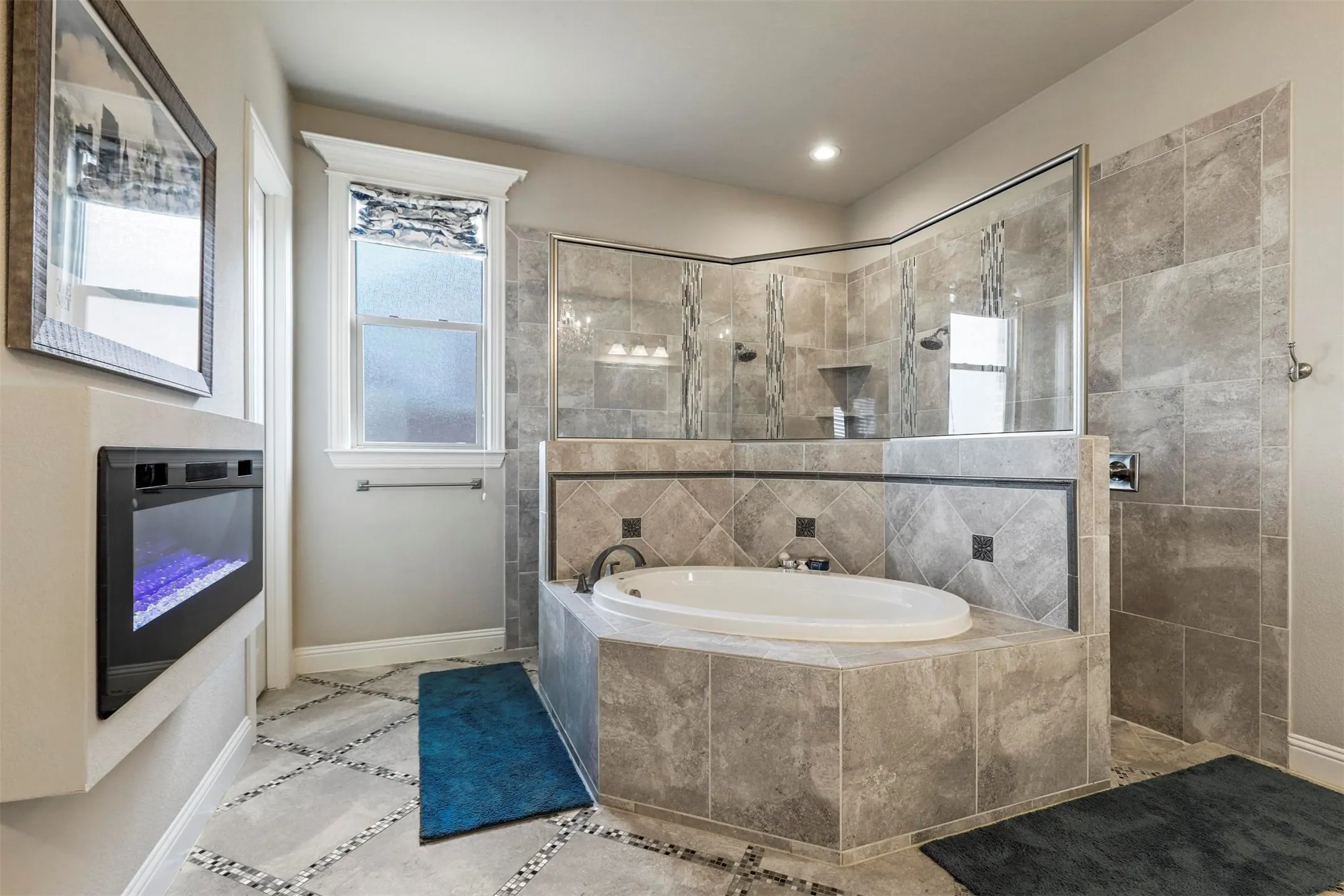 Soaking tub with surrounding stone-finish tile, glass-enclosed shower with matching tile, built-in electric fireplace, and patterned stone-finish floor