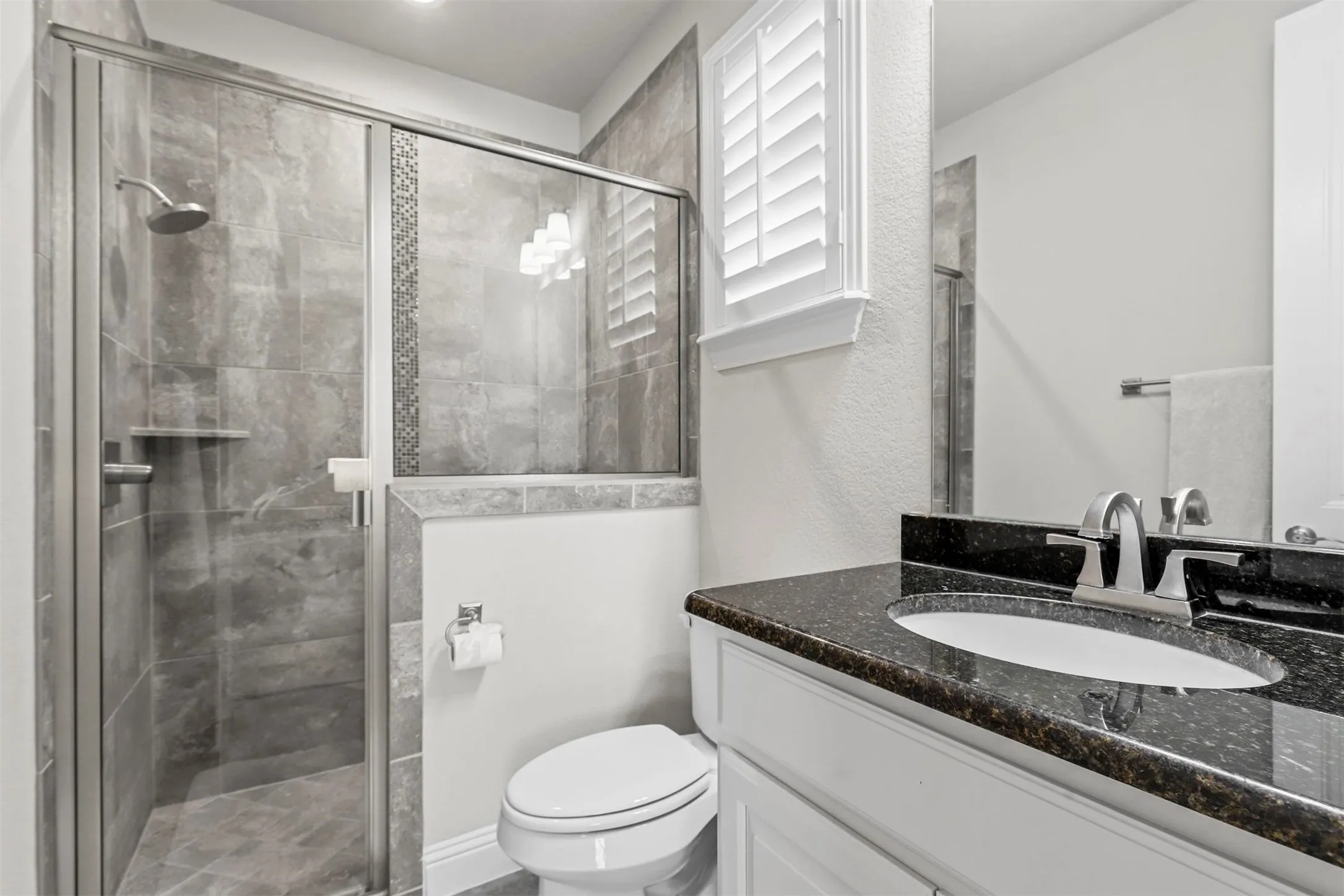 Walk-in shower with tile surround and glass enclosure, dark speckled countertop, white vanity with integrated sink, and white louvered window shutters
