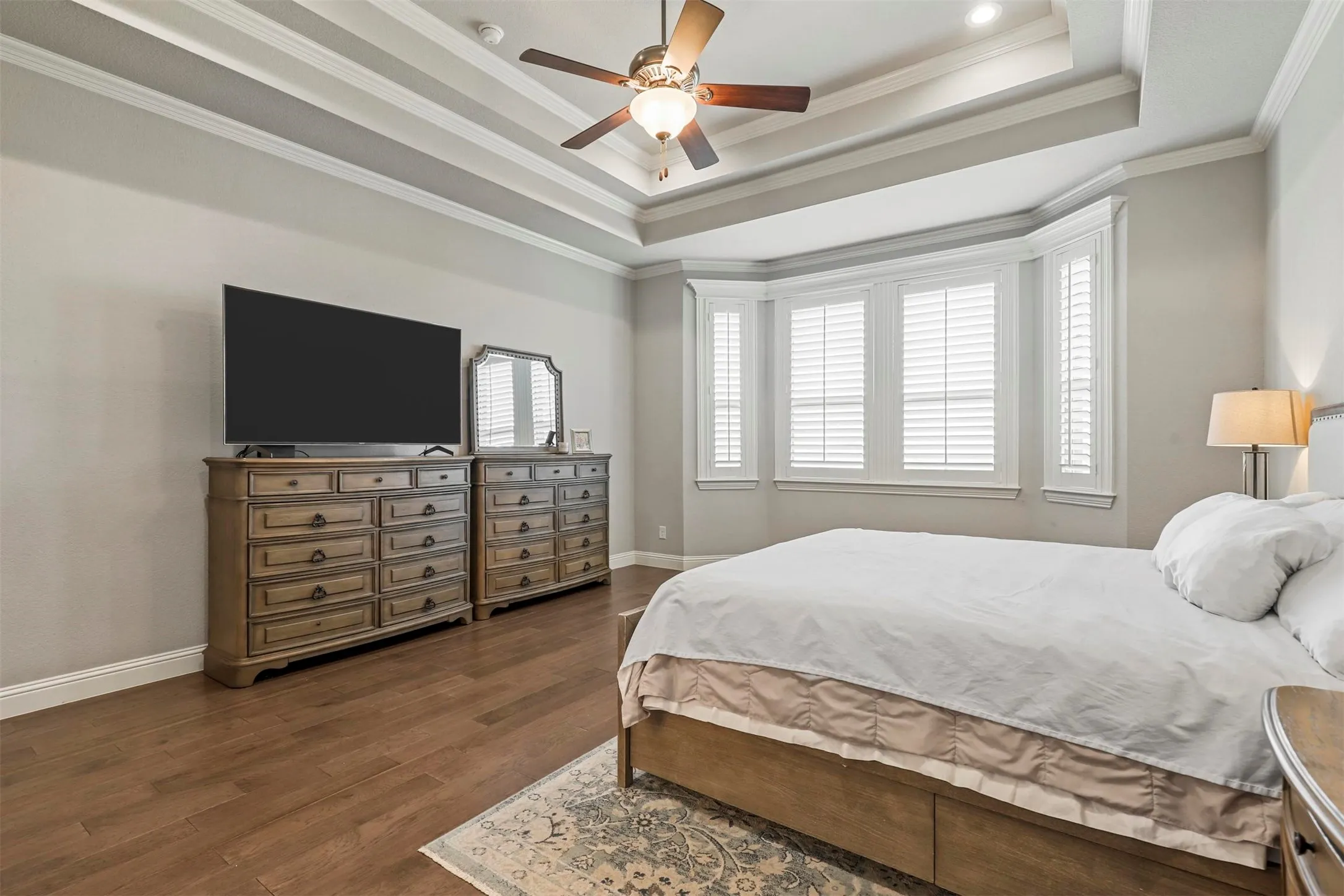 Tray ceiling with crown molding, ceiling fan, wood-finish flooring, bay window with plantation shutters, and recessed lighting