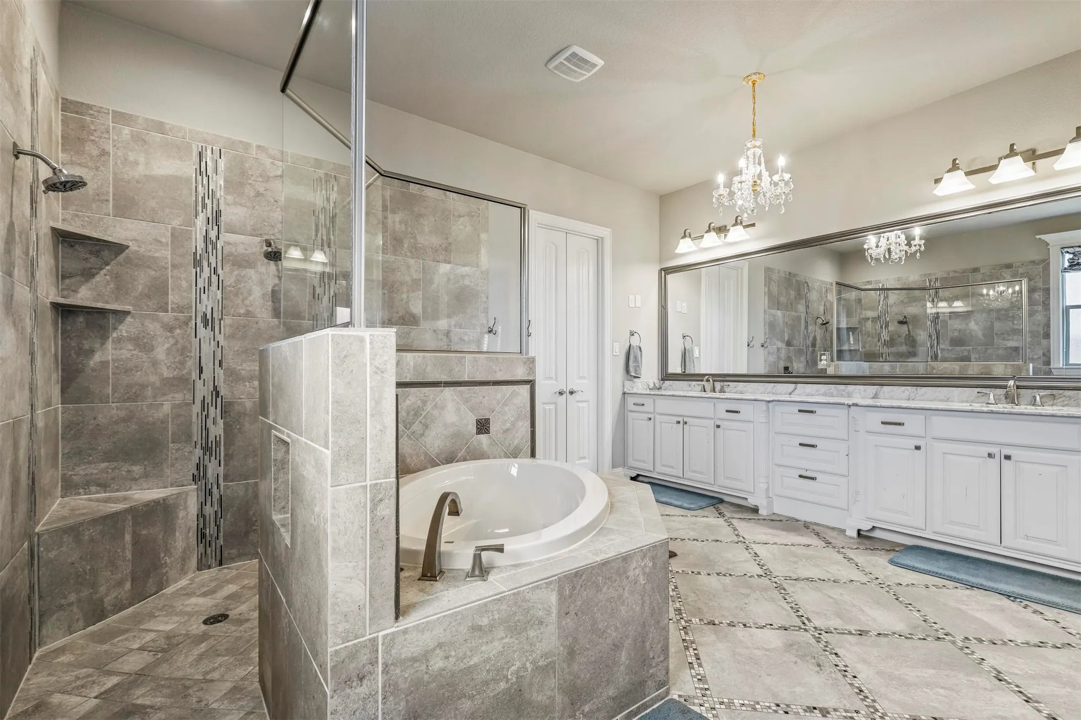 Spacious bathroom featuring a built-in soaking tub, a large tiled shower with built-in shelving, an extended vanity with dual sinks, and a decorative chandelier