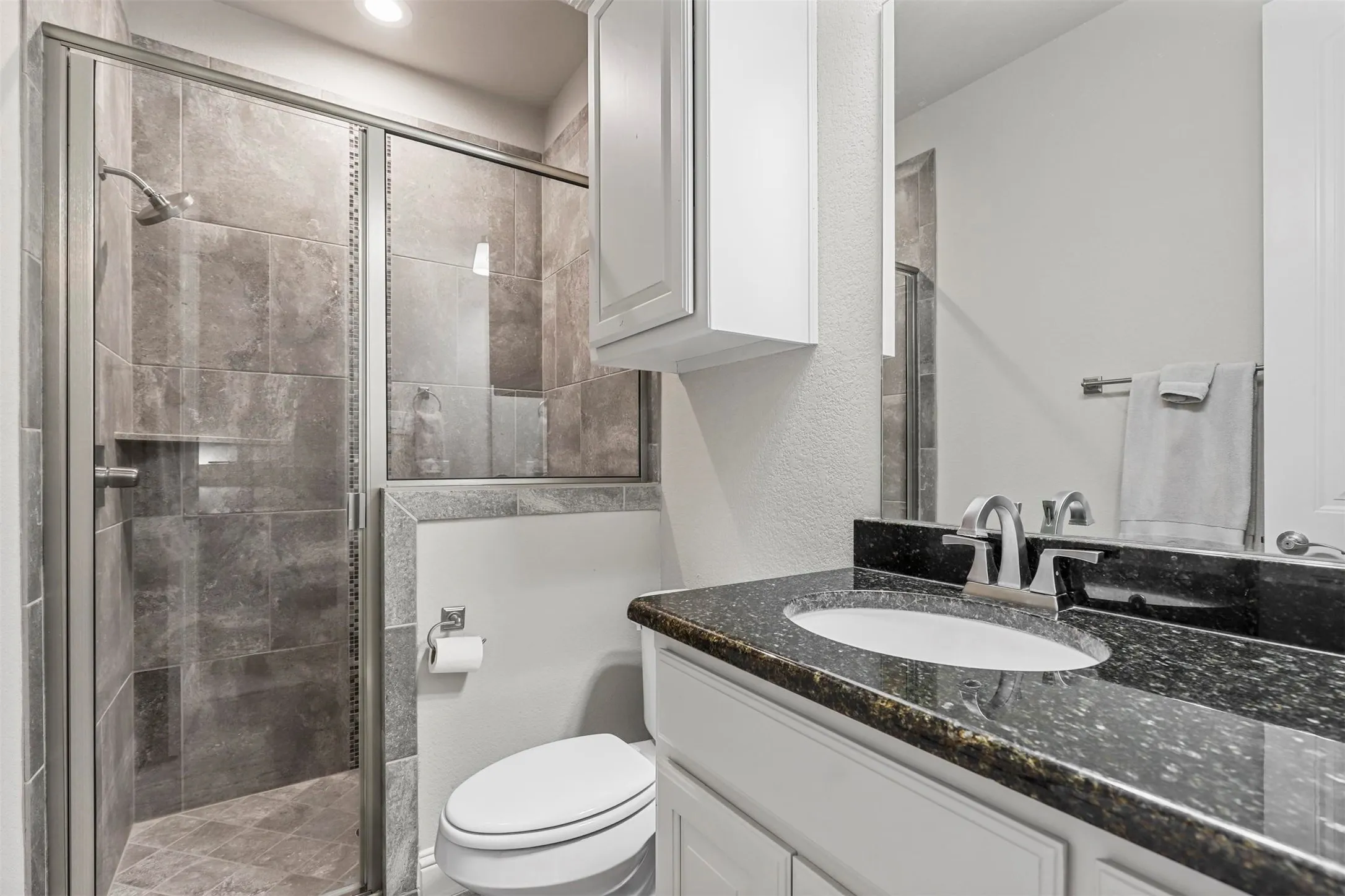Walk-in shower with tile surround and glass enclosure, vanity with stone countertop, under-mount sink, and white cabinetry