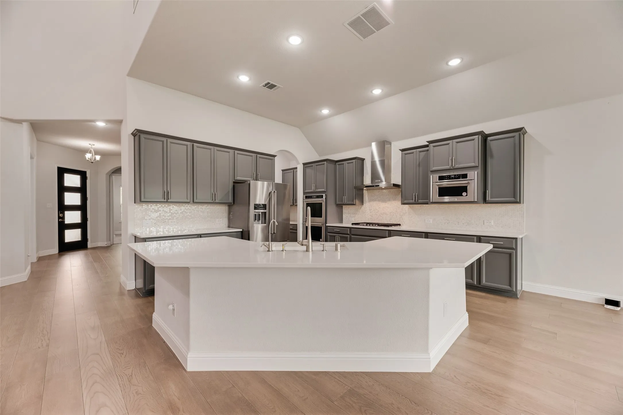 Spacious kitchen featuring an extended island with a built-in sink, stainless steel appliances, grey cabinetry, light-colored countertops, and wood-finish flooring