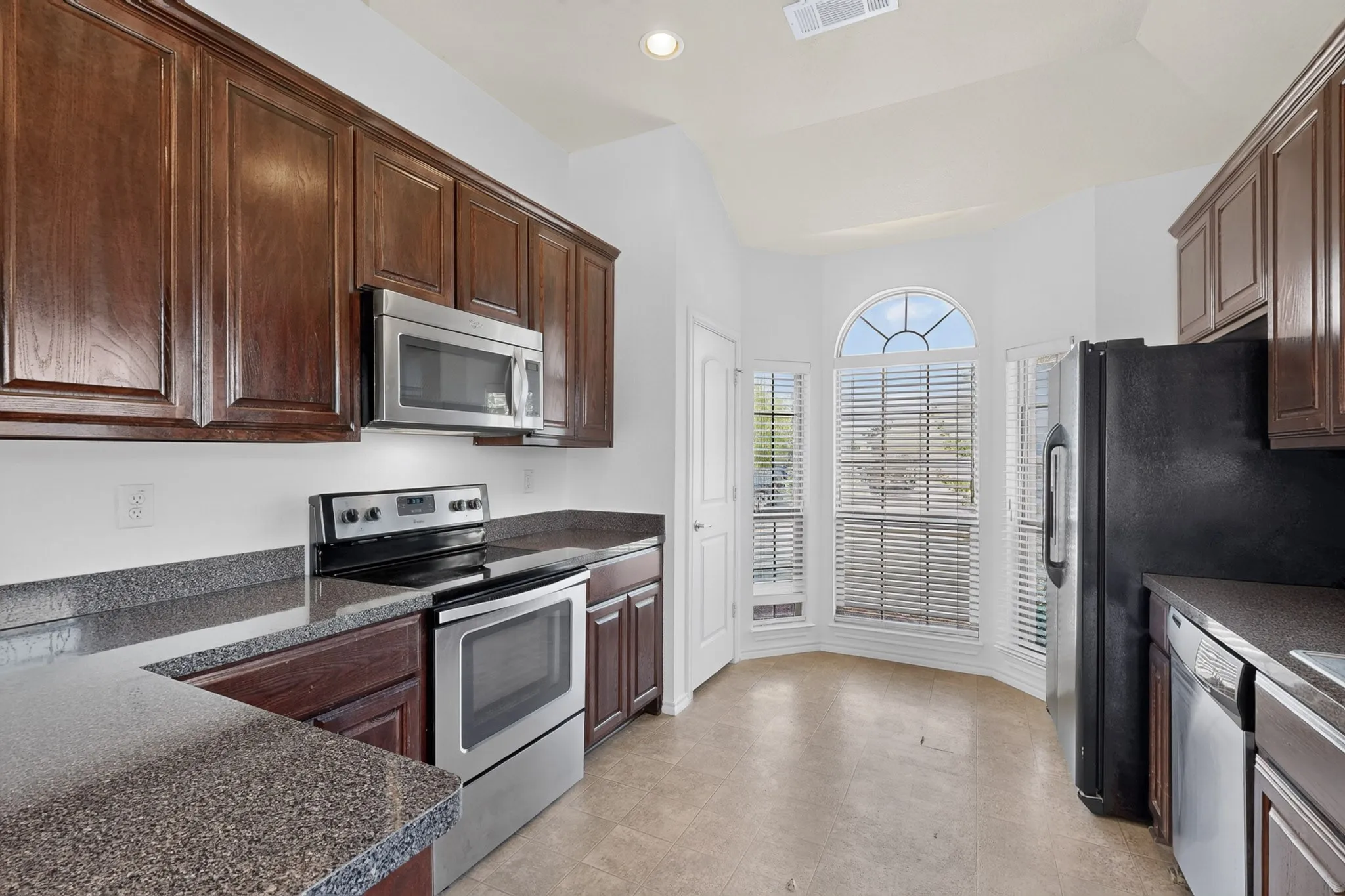 Kitchen featuring dark wood cabinetry, speckled countertops, stainless steel appliances, and a bay window with an arched transom