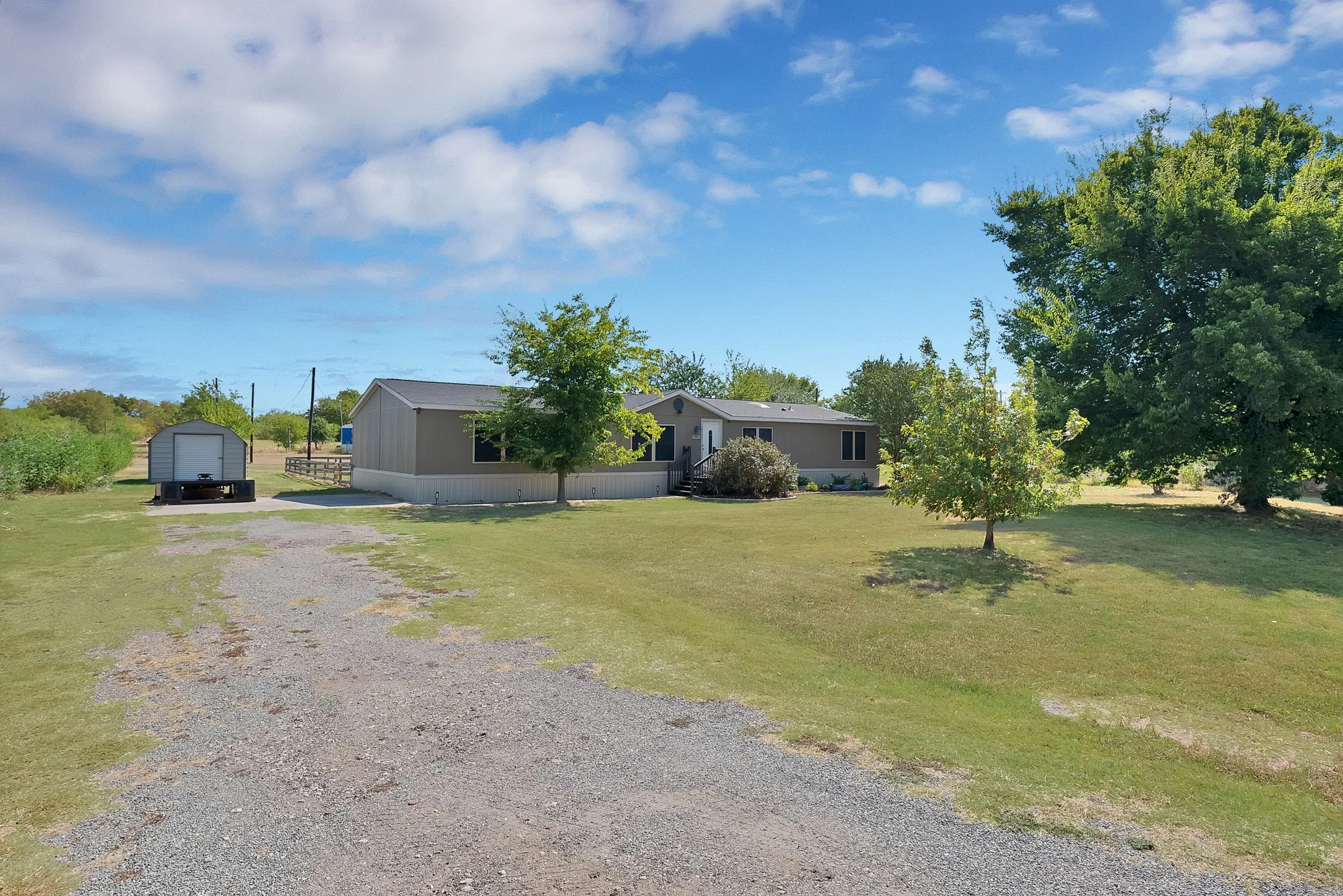 Expansive gravel driveway, detached storage shed, residential structure with neutral siding, mature trees, and a substantial grassy area
