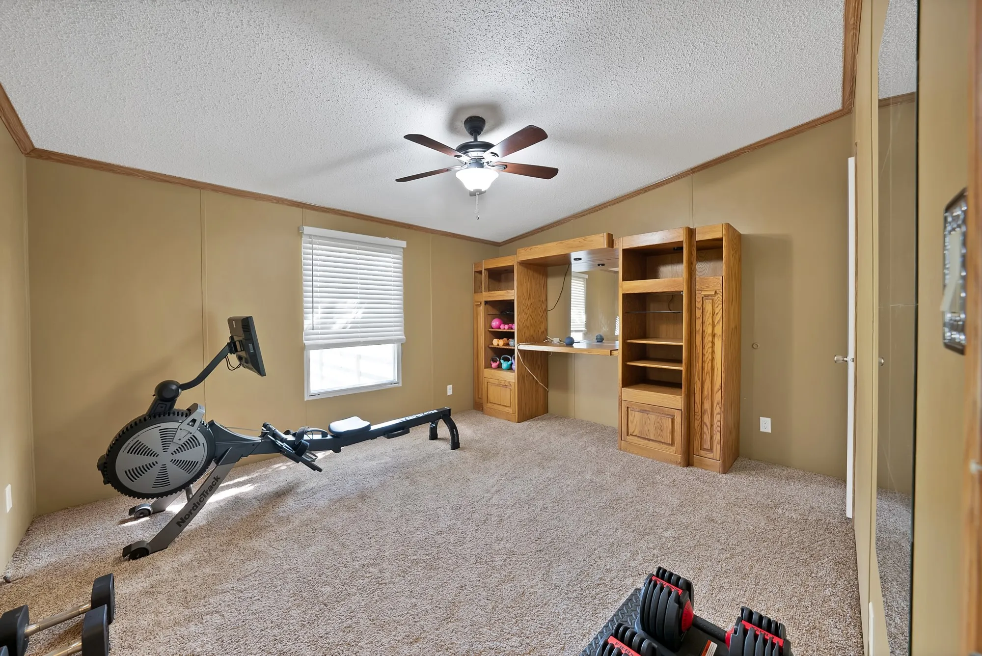 Carpeted room featuring a ceiling fan, window with blinds, and built-in wood cabinetry with shelving and a desk surface