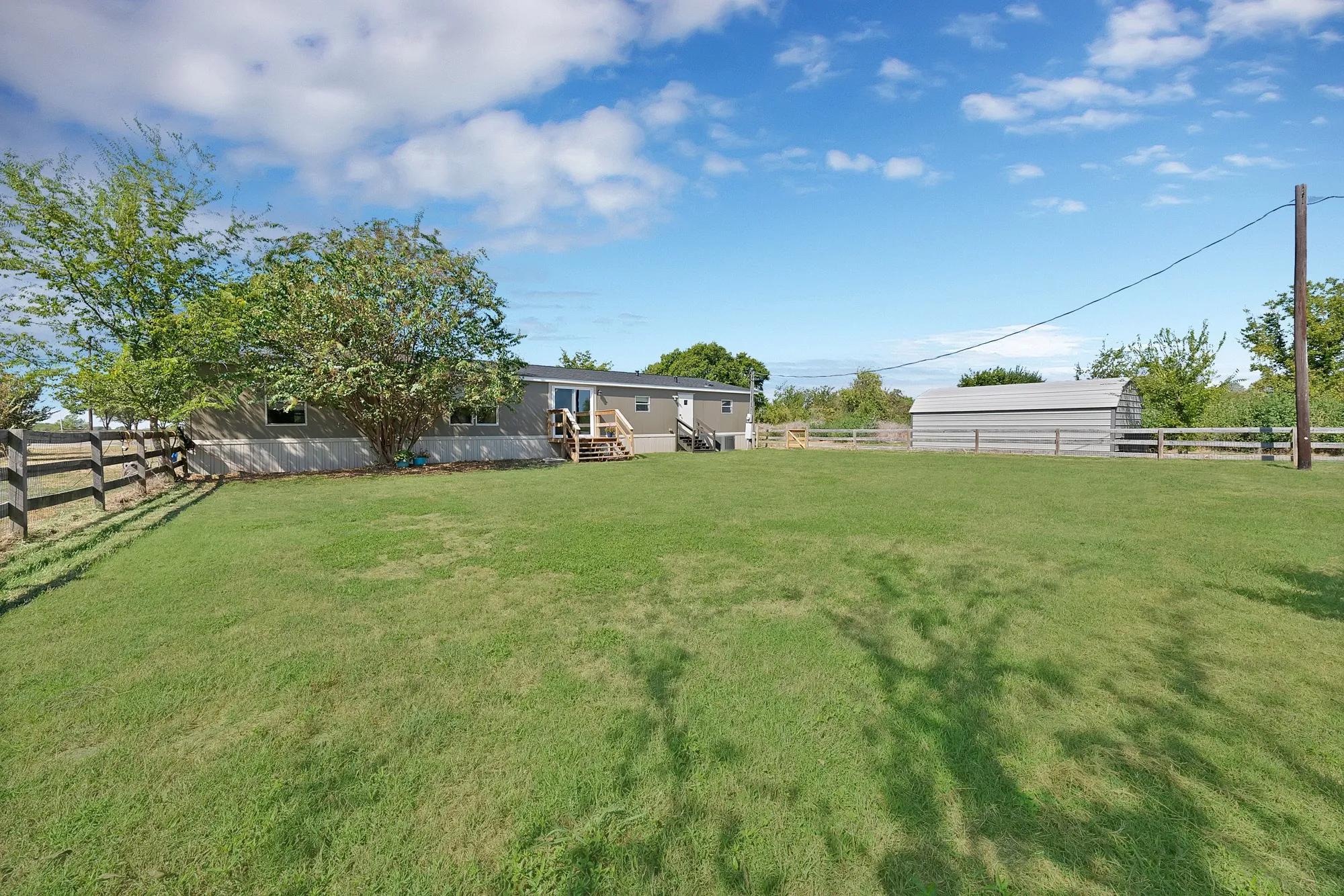 Expansive grassy yard with a split-rail fence, mature trees, and a detached building