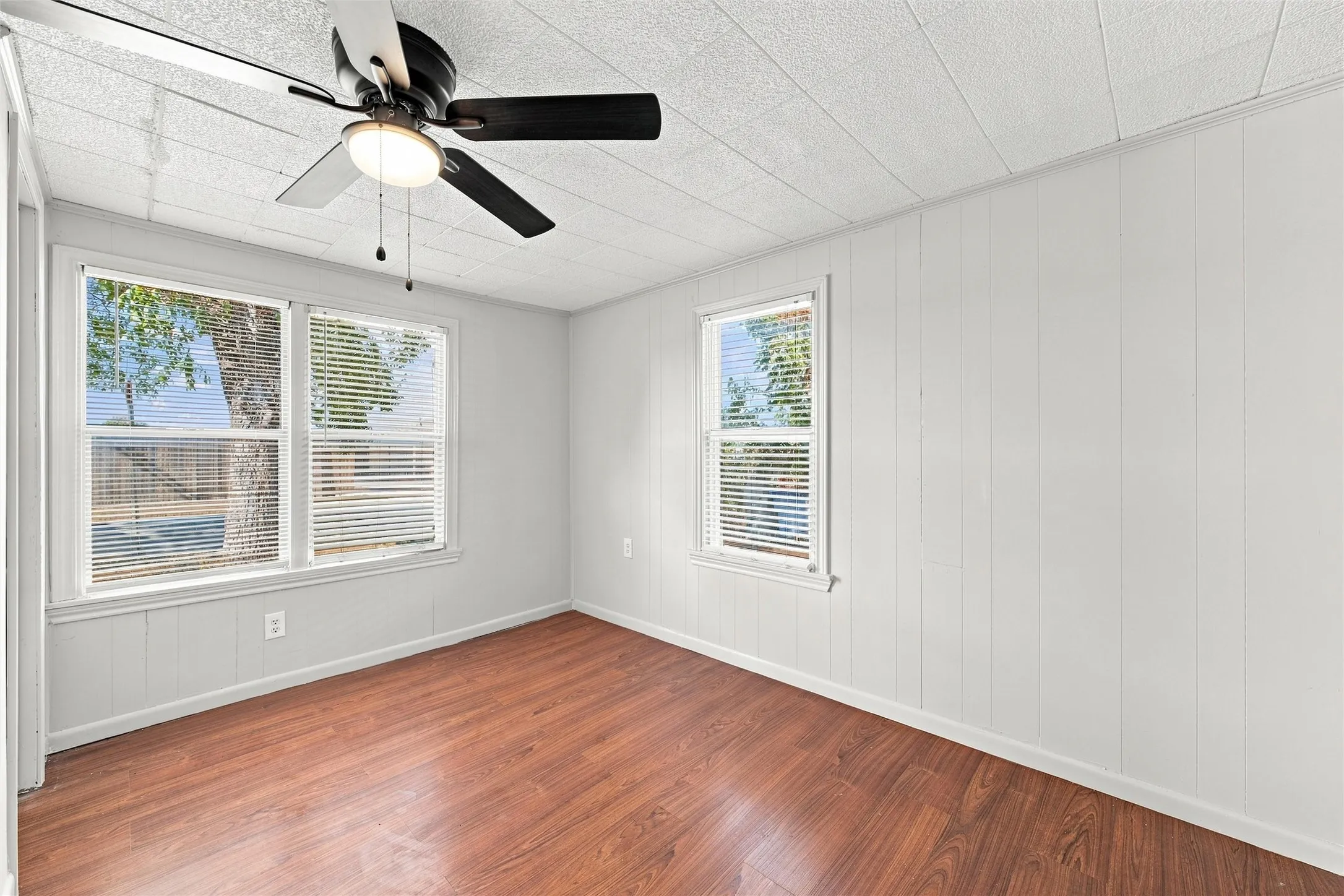 Wood-finish flooring, white paneled walls, ceiling fan with integrated lighting, multiple windows with blinds, and a textured ceiling