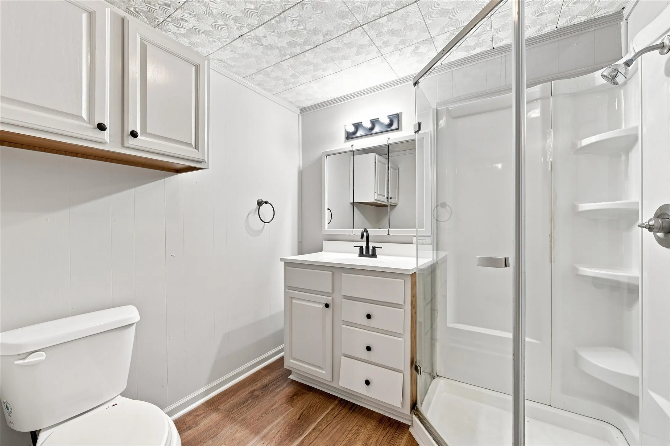 White vanity with black hardware, wood-finish flooring, and a walk-in shower with clear glass enclosure