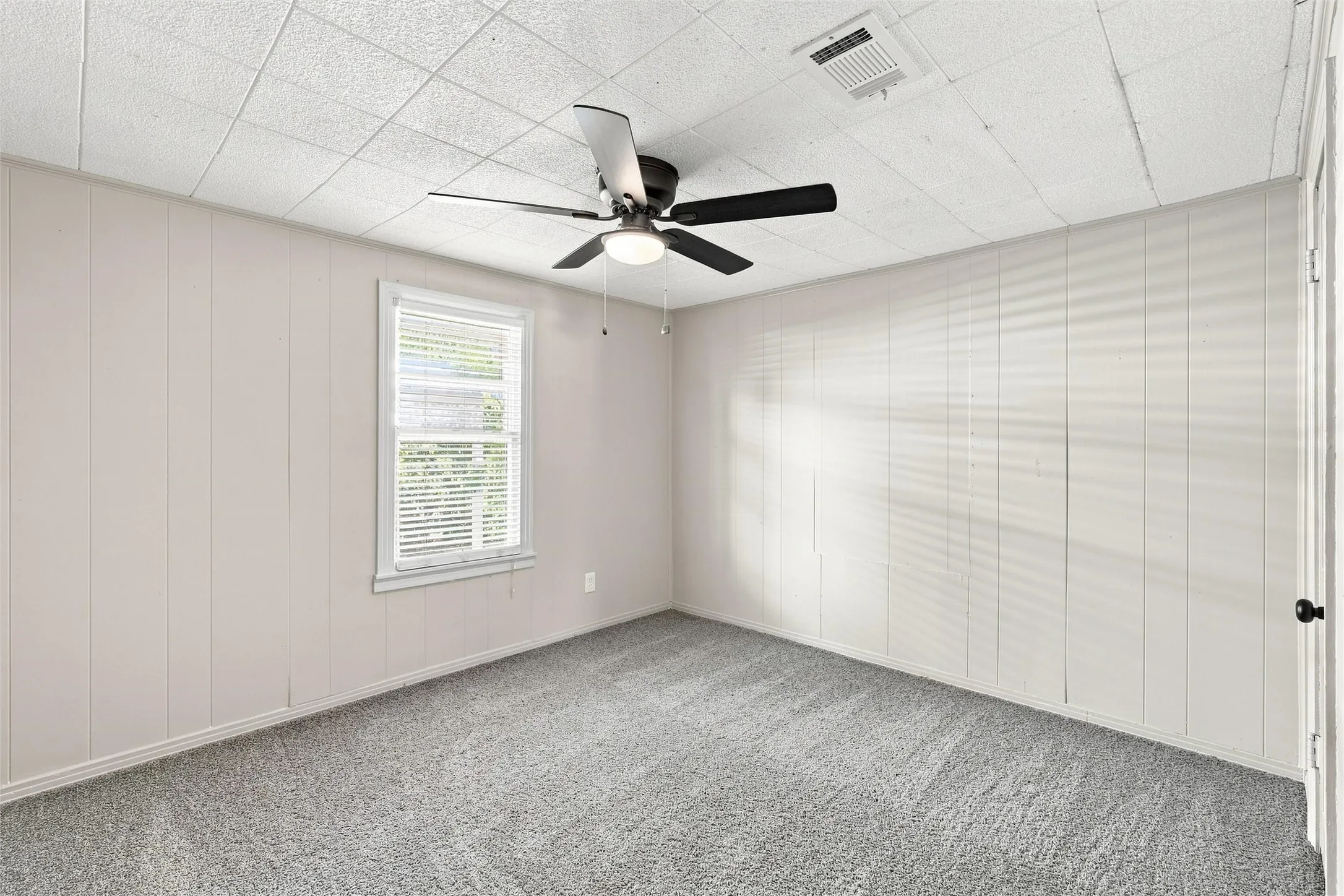 Carpeted flooring, paneled walls, ceiling fan with integrated lighting, and a white-framed window with blinds