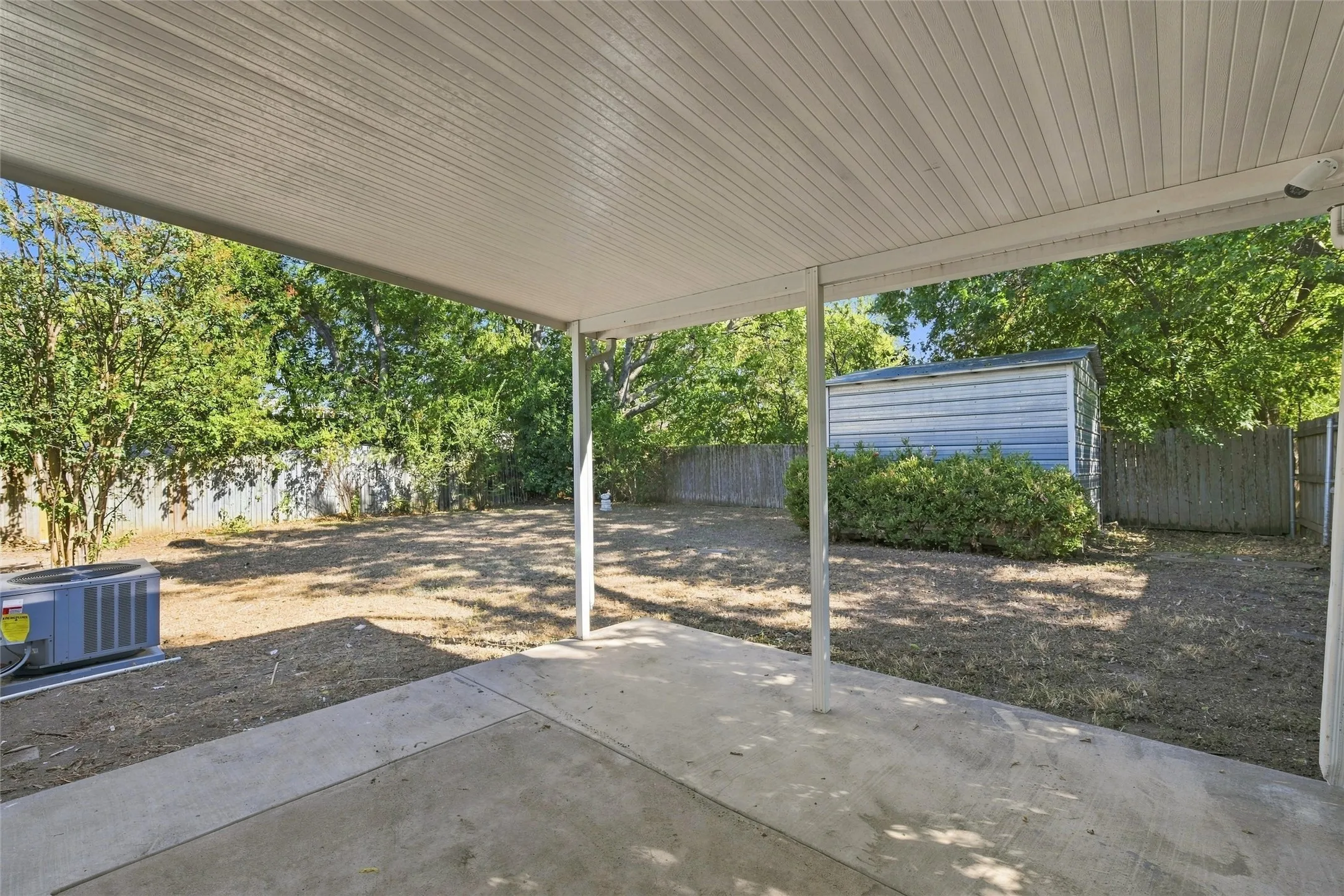 Covered patio with a concrete surface, fenced backyard, mature trees, and a storage shed