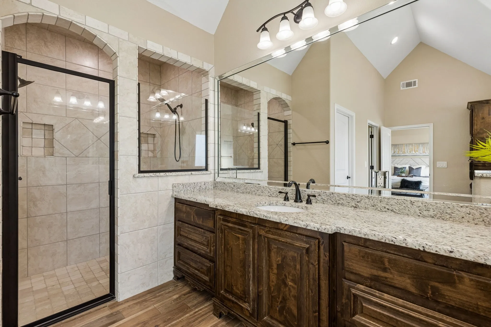 Vaulted ceiling with an expansive vanity mirror, dual-sink vanity with stone countertop, dark wood-finish cabinetry, and a walk-in shower with tiled walls