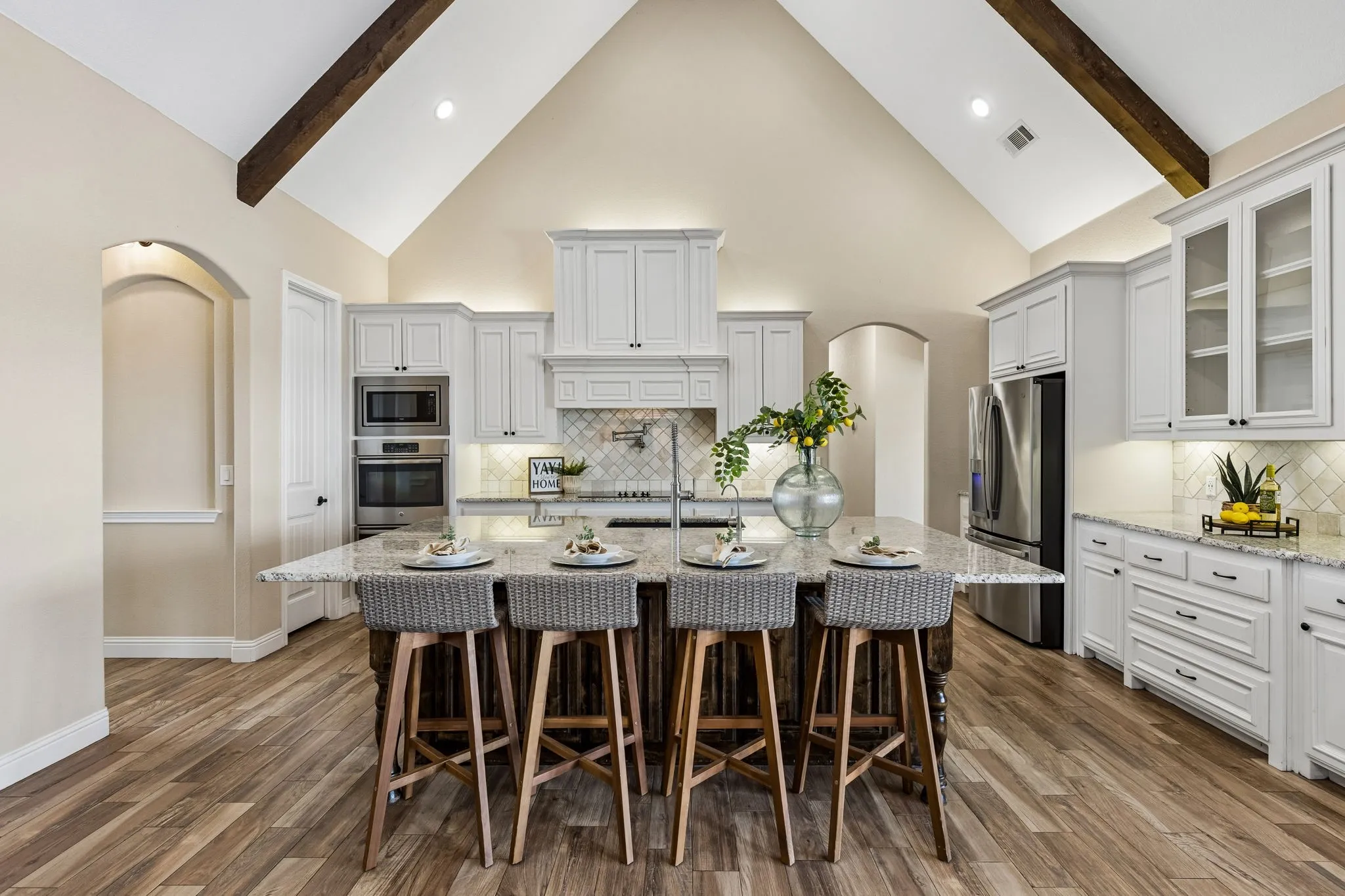 Vaulted ceiling with exposed wood beams, wood-finish flooring, white cabinetry with dark hardware, stainless steel appliances, and a central island with a stone-finish countertop