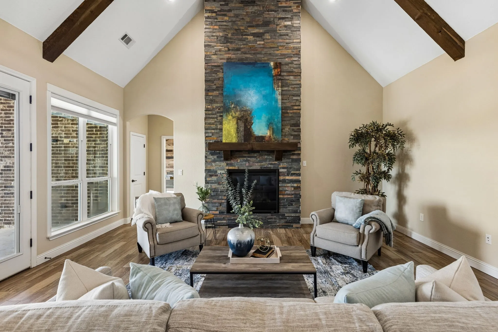 Vaulted ceiling with exposed wood beams, stone-clad fireplace with wood mantel, wood-finish flooring, and large windows with integrated blinds