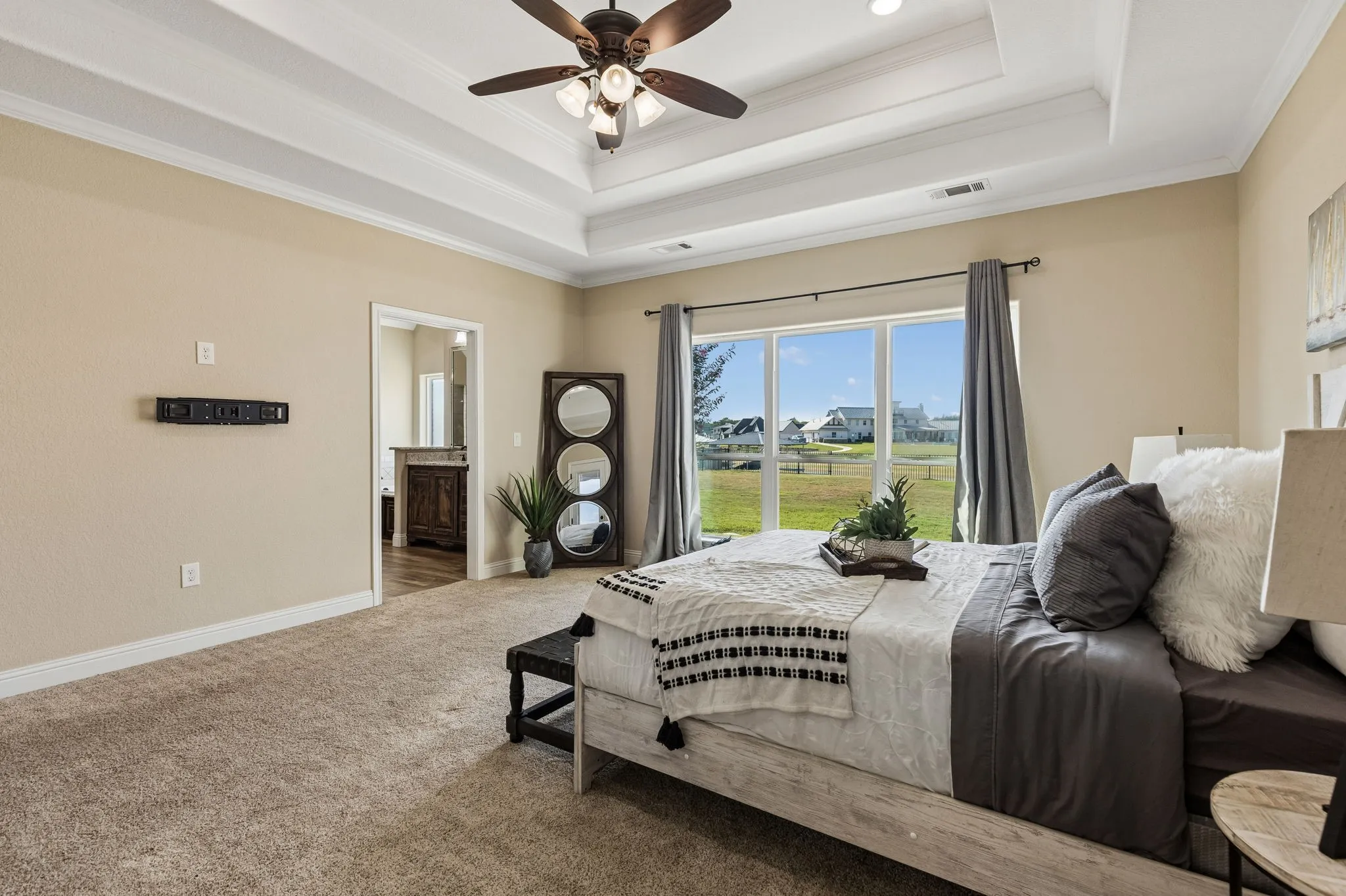 Tray ceiling with recessed lighting and a ceiling fan, broad window with outdoor scenery, wall-to-wall carpeting, and an open doorway to an adjacent space with wood-finish flooring and a built-in vanity