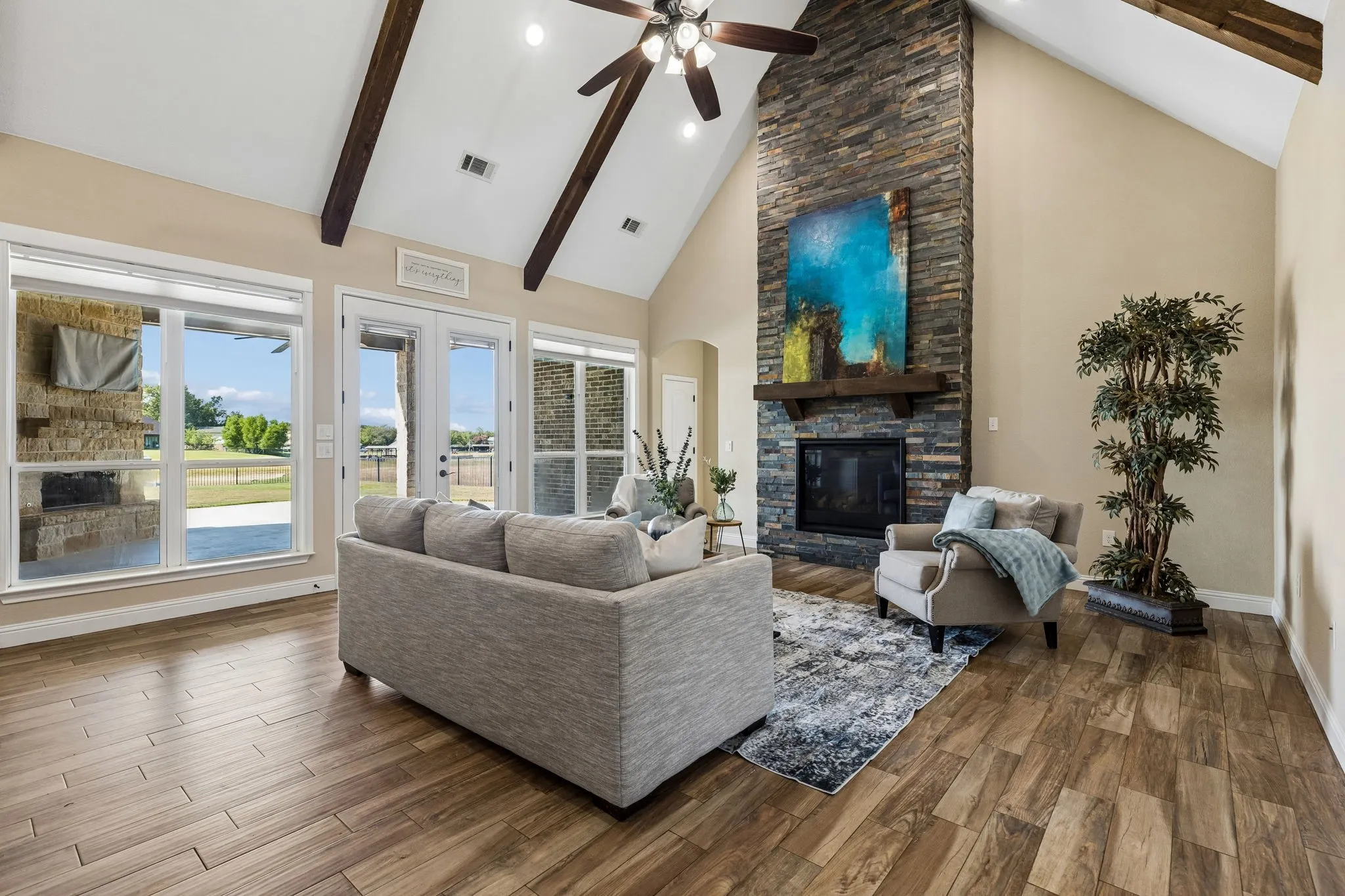 Vaulted ceiling with exposed wood beams, wood-finish flooring, stone-clad fireplace, ceiling fan, and ample natural light through large windows