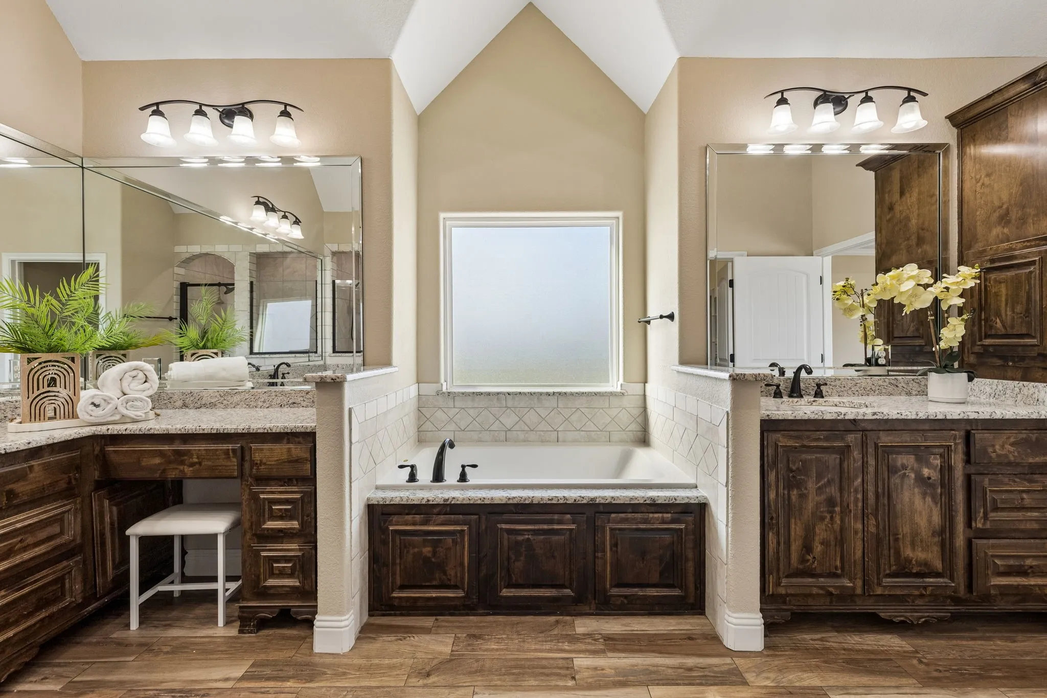 Dual vanities with stone countertops, wood-finish flooring, a built-in tub with tile surround, and vaulted ceilings