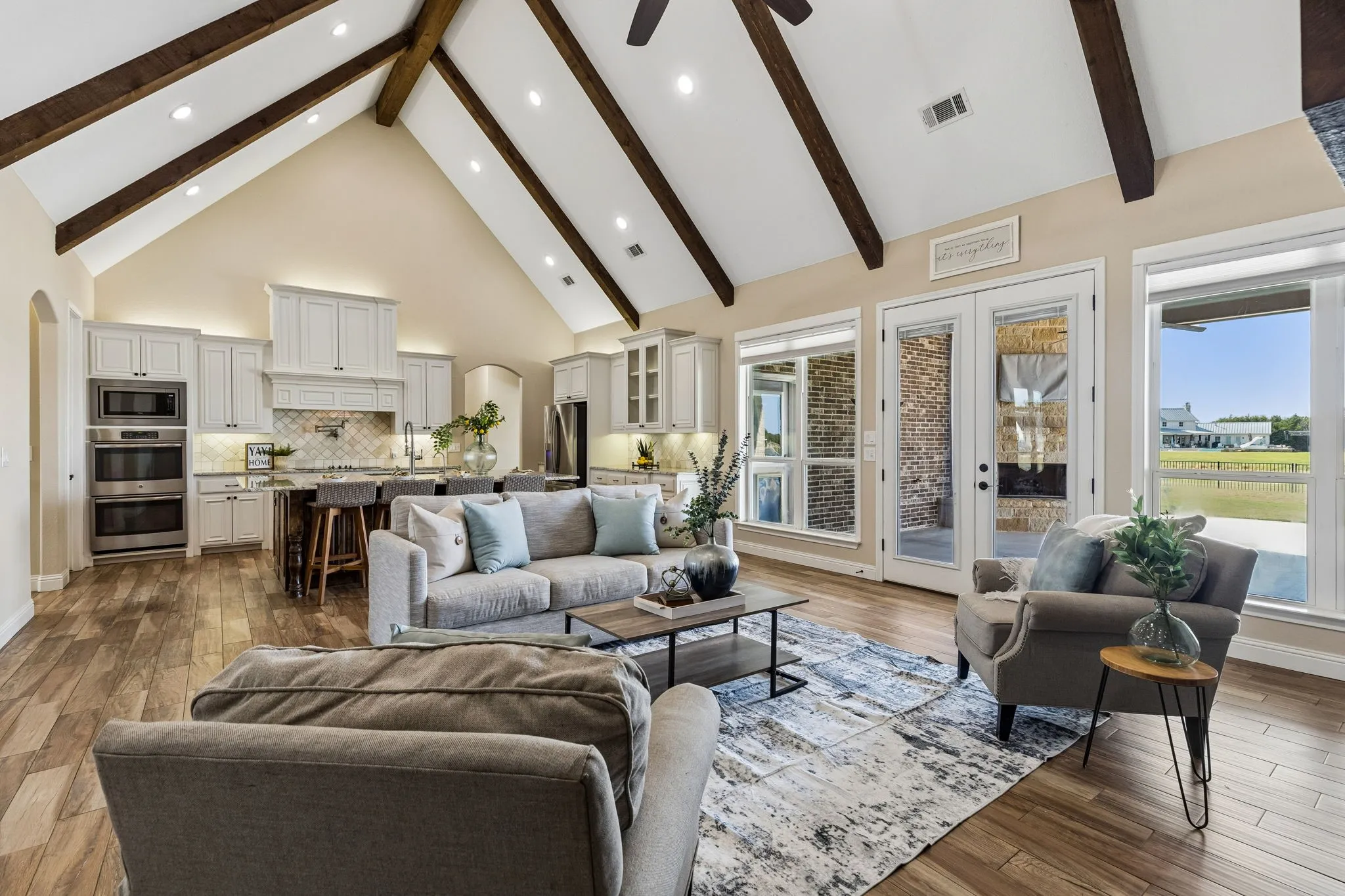 Vaulted ceiling with exposed wood beams, wood-finish flooring, white cabinetry, built-in stainless steel double ovens, and double glass-paneled doors