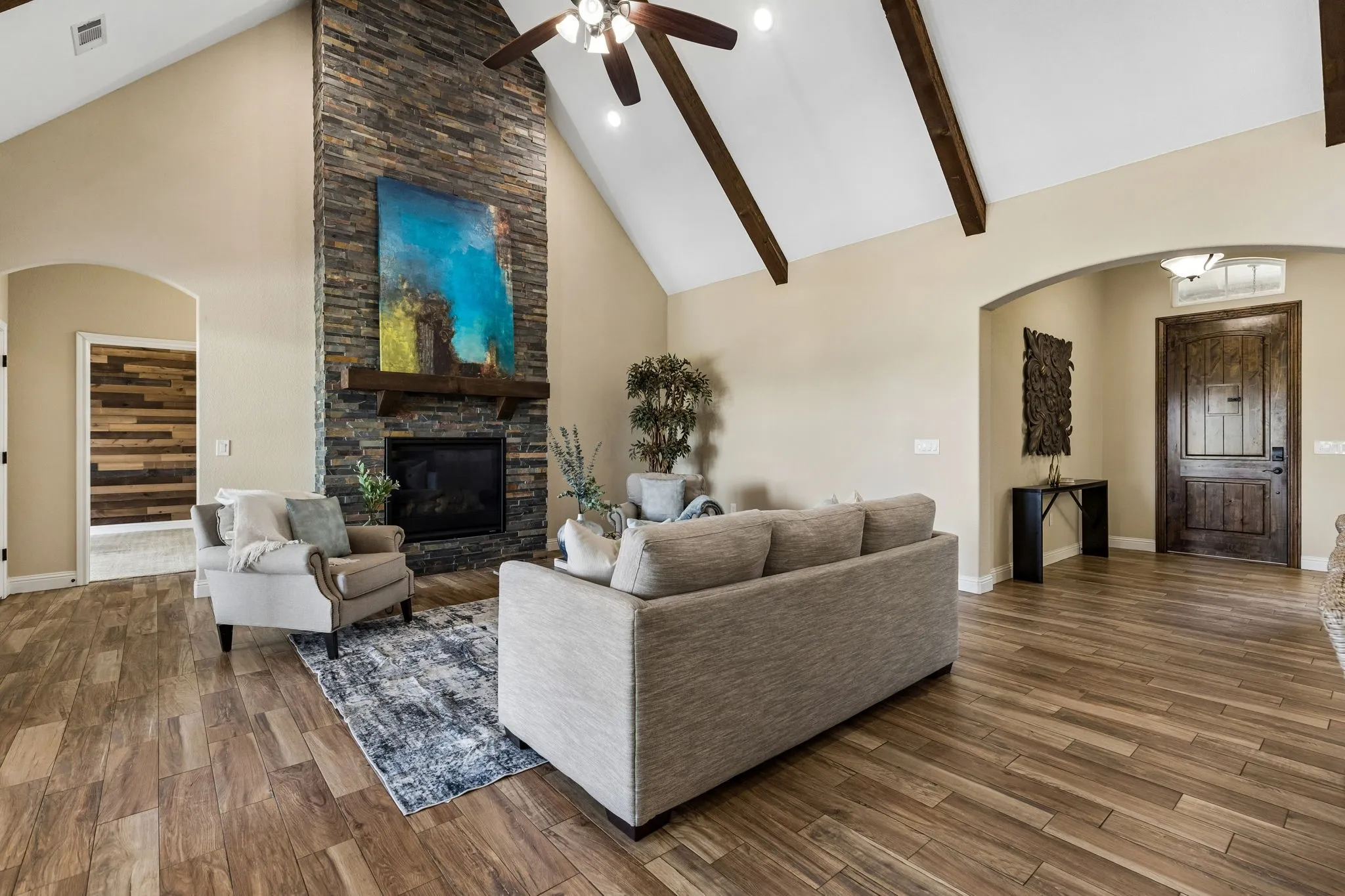 Vaulted ceiling with exposed wood beams, wood-finish flooring, stone-clad fireplace with wood mantel, arched doorways, and a dark wood entry door